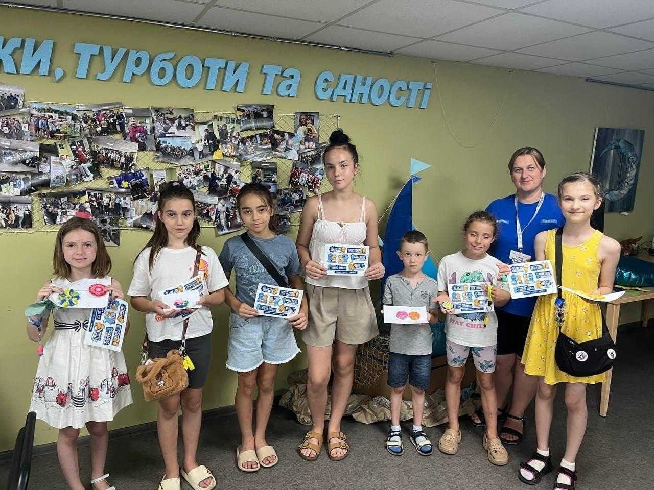 Учасники дитячого майстер-класу в дніпровському хабі «Вільні разом» тримають у руках свої розмальовані гіпсові фігурки та дипломи. Сім хлопчиків та дівчат різного віку стоять у ряд поруч із представницею БФ «Посмішка» у синій футболці.