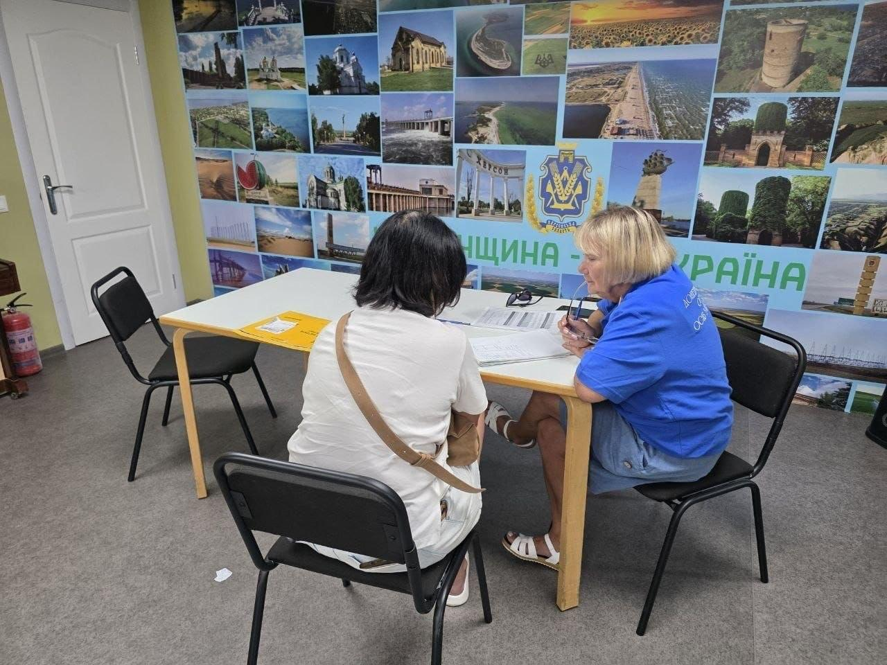 Юридична та соціальна консультація переселенки з Херсонщини в координаційному центрі «Вільні разом» у Дніпрі. Юрисконсульт ГО «Аваліст» та кейс-менеджер спілкуються з відвідувачкою за робочим столом. На задньому плані — фотостіна з краєвидами та написом «Херсонщина — це Україна».