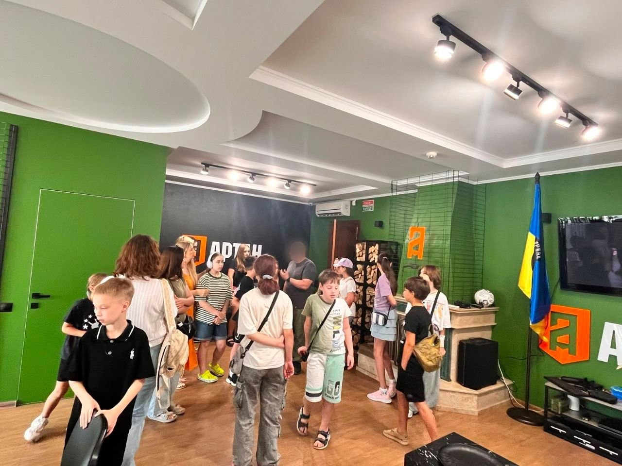 Екскурсія для дітей-переселенців із Херсонщини простором спецпідрозділу ГУР МО «Артан» у Миколаєві. Група підлітків оглядає зал та спілкується з інструктором, обличчя якого приховане розмиттям. Зустріч заходу «Активна молодь – сильна громада» проходить у кімнаті із зеленими стінами, прапором України та символікою підрозділу.