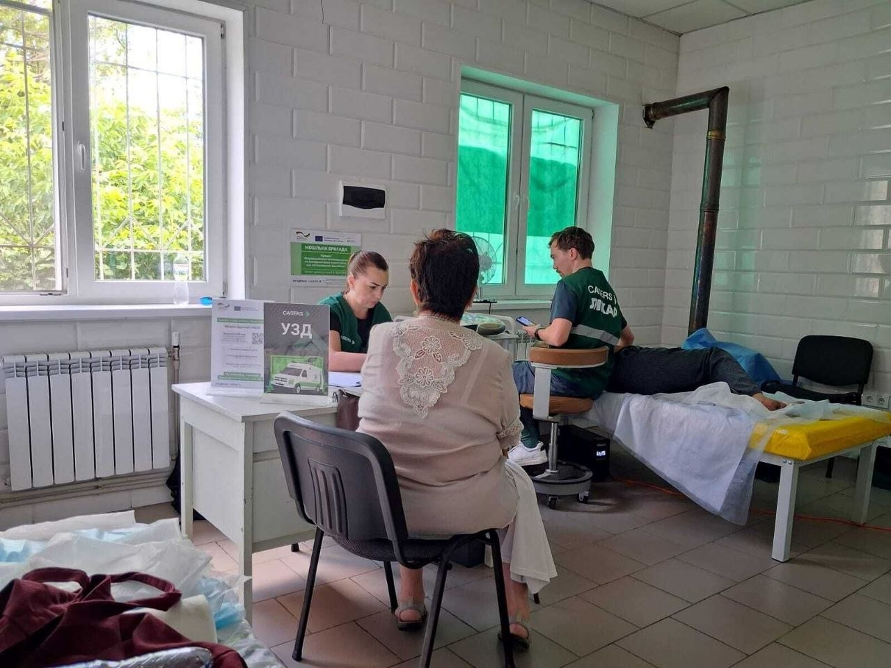 Кардіохірург Інституту ім. М.М. Амосова Святослав Калашніков проводить УЗД серця пацієнту на канапі, а медсестра за столом із табличкою «УЗД» консультує іншу жінку. Візит мобільної бригади «CASERS» організовано для мешканців Чорнобаївки та Калинівського в кабінеті місцевого закладу охорони здоров'я.