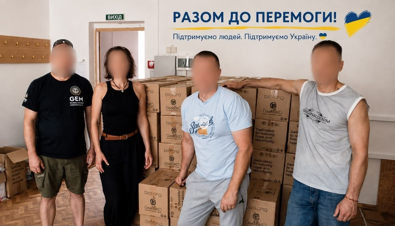 Чотири особи з розмитими обличчями стоять біля великої кількості коробок із продуктовими наборами для родин із лівобережжя Херсонщини. Ліворуч стоїть представник організації GEM у брендованій чорній футболці та жінка в чорному одязі, а праворуч — два чоловіки у світлих футболках. На білій стіні над допомогою від фондів GEM та HGBF розміщено великий напис «РАЗОМ ДО ПЕРЕМОГИ! Підтримуємо людей. Підтримуємо Україну.» із синьо-жовтими символами.