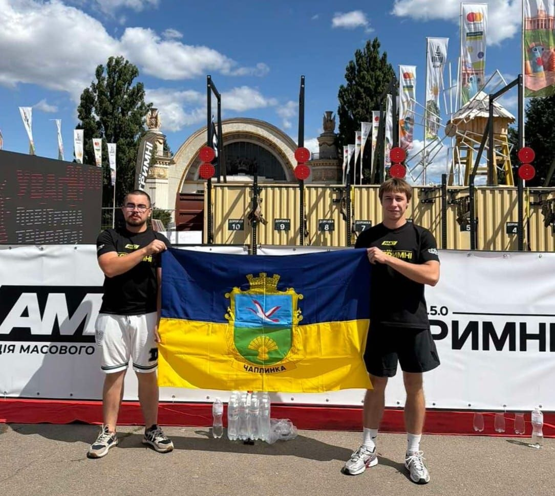 Два спортсмени з Чаплинської громади тримають прапор селища Чаплинка під час спортивно-патріотичного заходу Героїчний комплекс «Павук» пам’яті Героя України Богдана Карватка у Києві. Хлопці у чорних футболках стоять на тлі спортивних майданчиків під відкритим небом та білих банерів Агенції масового спорту України.