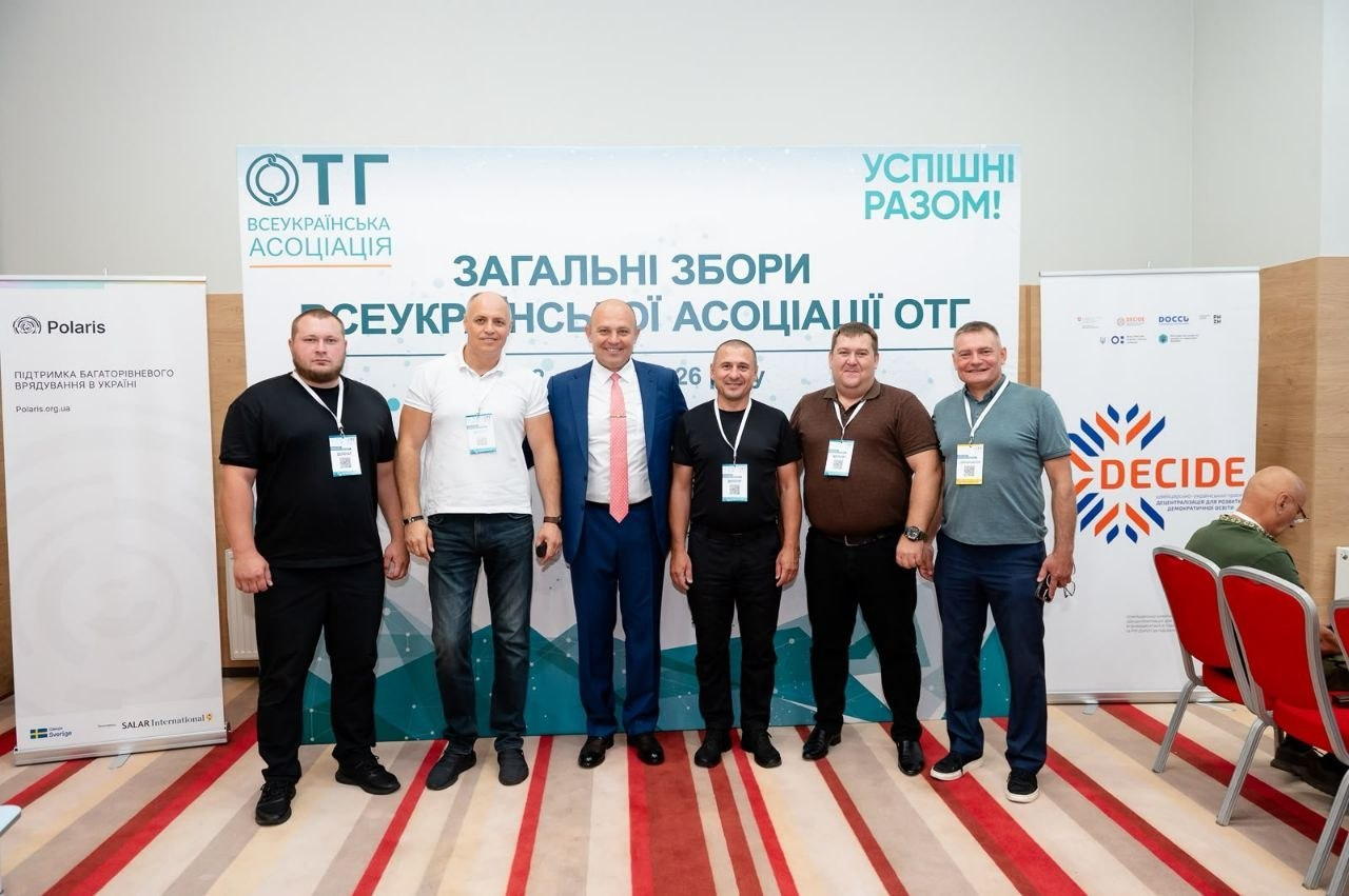 Делегація представників територіальних громад Херсонщини стоїть у ряд на Загальних зборах Всеукраїнської асоціації ОТГ у Києві. Шість чоловіків посміхаються на тлі головного білого банера заходу з написами «Загальні збори» та гаслом «Успішні разом!». По обидва боки розміщено банери партнерів проєкту — «Polaris» (ліворуч) та «DECIDE» (праворуч).