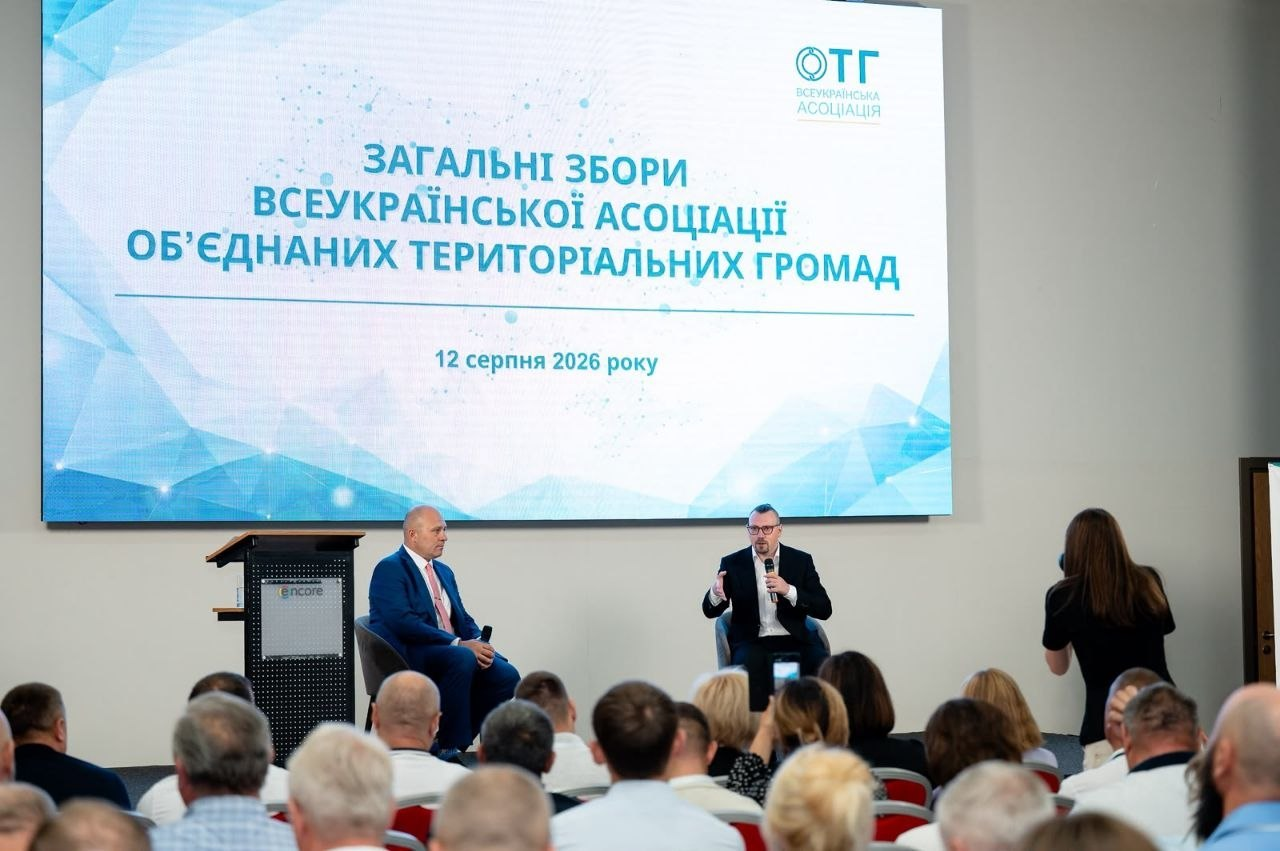 Сцена конференц-залу під час Загальних зборів Всеукраїнської асоціації об’єднаних територіальних громад 12 серпня 2026 року. На великому екрані відображено синьо-блакитну заставку заходу. У кріслах на сцені сидять двоє спікерів: ліворуч — переобраний голова Асоціації Олександр Корінний у синьому костюмі та червоній краватці, праворуч — Міністр Віталій Безгін у чорному піджаку, білій сорочці та окулярах, який виступає з мікрофоном перед залом, заповненим делегатами.
