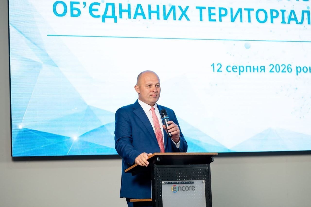 Голова Всеукраїнської асоціації ОТГ Олександр Корінний у синьому костюмі та червоній краватці виступає з мікрофоном за трибуною під час Загальних зборів асоціації. На великому мультимедійному екрані за його спиною видно частину заголовка та дату з'їзду — «12 серпня 2026 року».