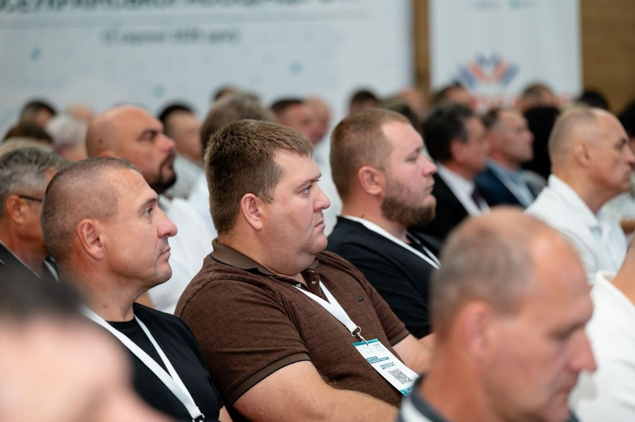Члени делегації Херсонщини сидять у залі серед інших учасників Загальних зборів Всеукраїнської асоціації ОТГ. На передньому плані зображені троє чоловіків у темних футболках та поло з офіційними білими бейджами «Делегат», які зосереджено слухають виступи спікерів. Решта зали та присутніх на з’їзді перебувають у розмитті.
