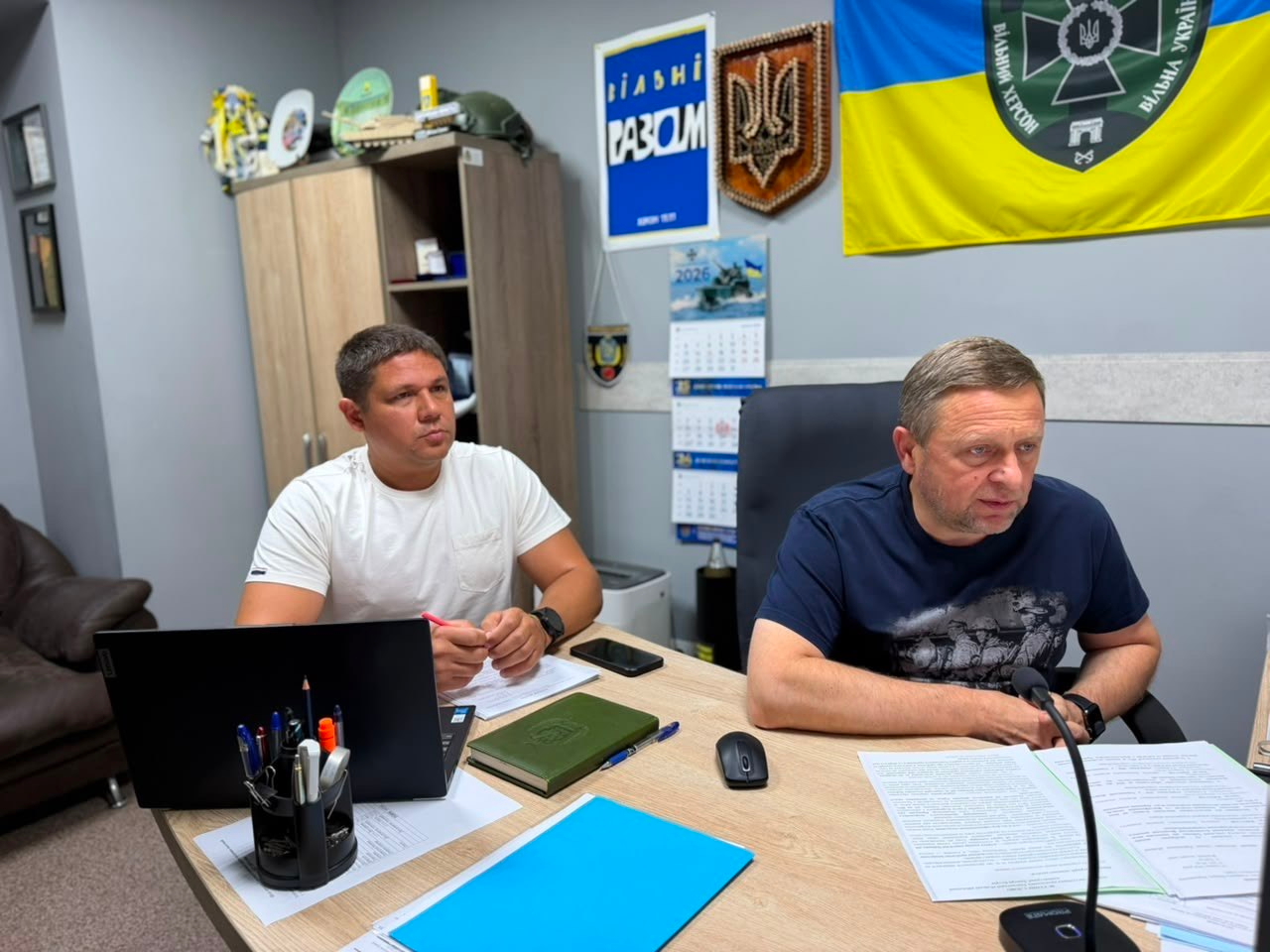 Перший заступник начальника Херсонської ОВА Дмитро Бутрій (по центру за столом у темно-синій футболці-поло) та директор Департаменту розвитку сільського господарства та зрошення ХОДА Дмитро Юнусов (ліворуч у білій футболці) проводять робочу онлайн-нараду. Вони вивчають документи та таблиці у робочому кабінеті на тлі державного прапора України. Праворуч на передньому плані видно великий комп'ютерний монітор.