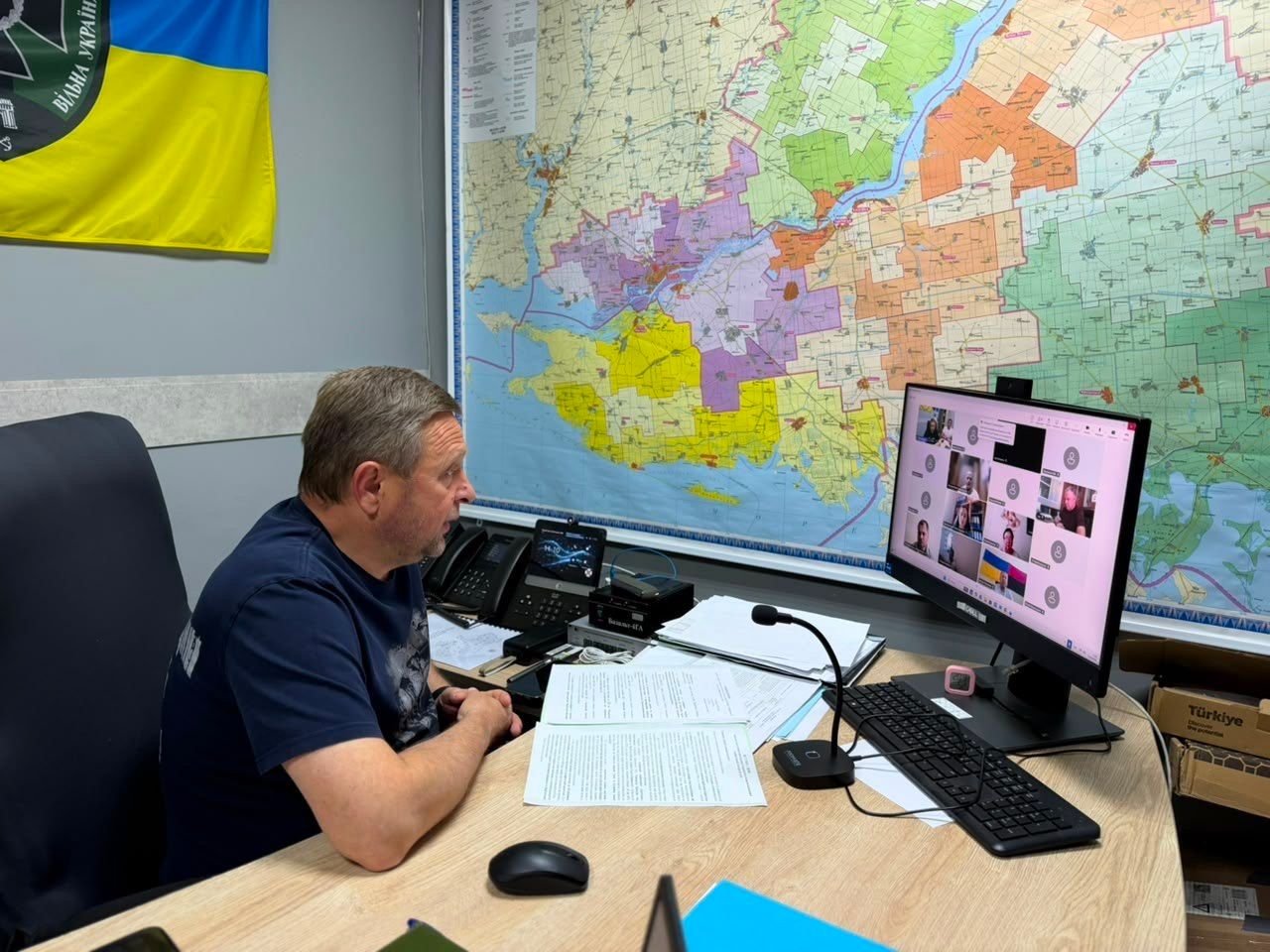 Перший заступник начальника Херсонської ОВА Дмитро Бутрій бере участь в онлайн-нараді з аграріями Херсонщини. Посадовець у темно-синій футболці сидить у профілі за робочим столом перед монітором із вікнами учасників у Zoom. На стіні за ним розміщена велика кольорова мапа Херсонської області та державний прапор України із символікою.
