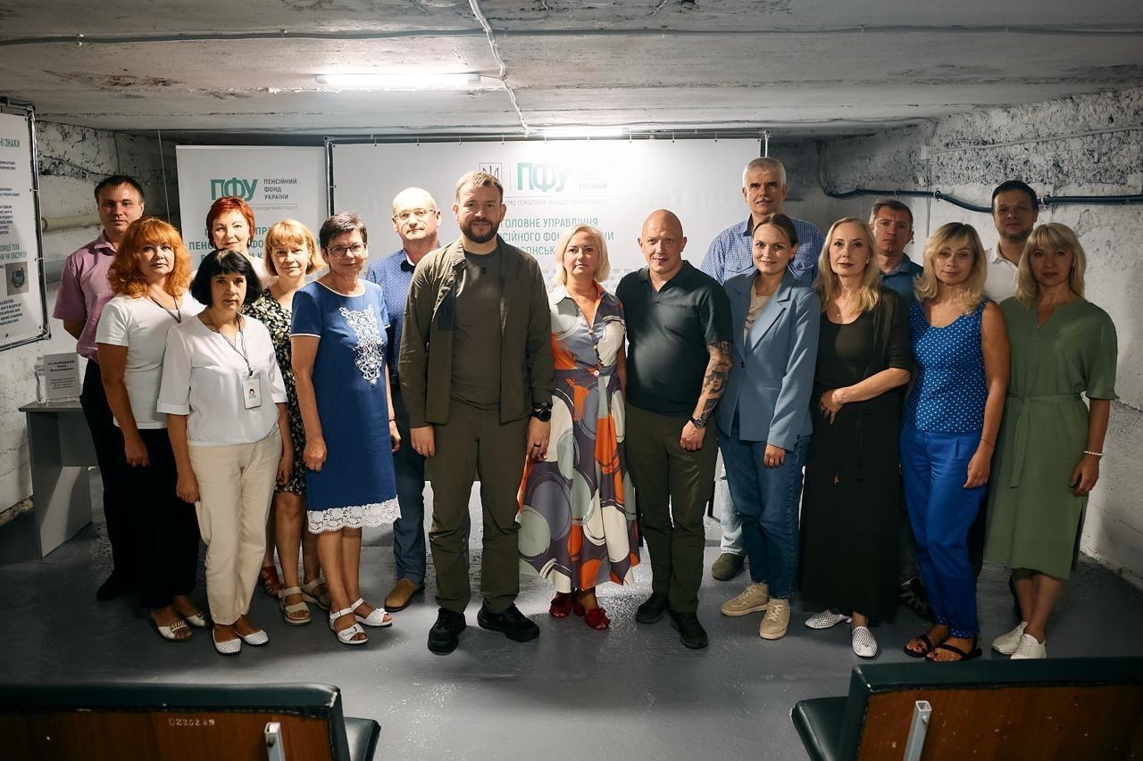 Групове фото Міністра соціальної політики, сім’ї та єдності України Дениса Улютіна, заступника начальника Херсонської ОВА Євгенія Ігнатенка та колективу Головного управління Пенсійного фонду України в Херсонській області. Група людей стоять у підвальному приміщенні (укритті) на тлі офіційного інформаційного банера установи. У центрі першого ряду стоїть Денис Улютін у військово-зеленому одязі (хакі), а праворуч від нього — Євгеній Ігнатенко у темно-сірій футболці поло. Візит урядовця присвячений ознайомленню з роботою сервісного центру в умовах воєнного стану.