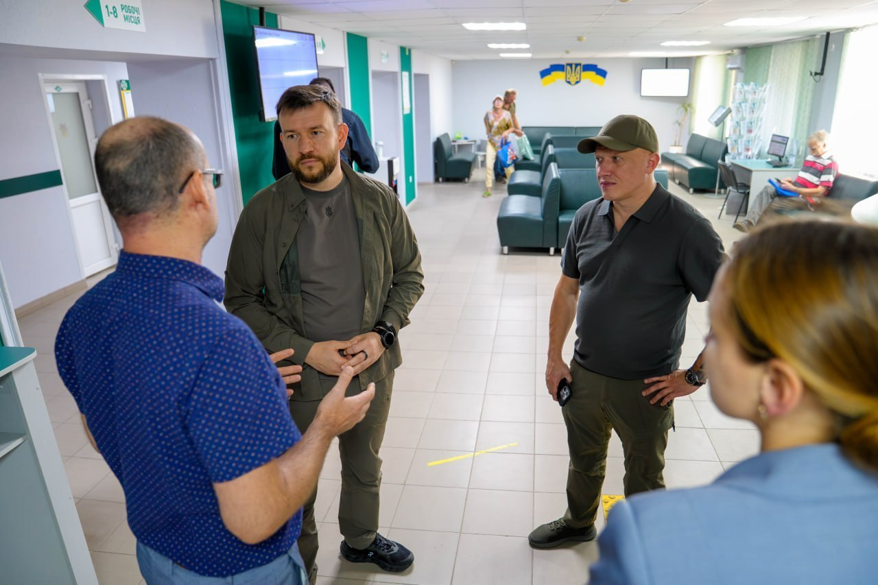 Міністр соціальної політики, сім’ї та єдності Денис Улютін у зеленому одязі та заступник начальника Херсонської ОВА Євгеній Ігнатенко у кепці та темно-сірому поло спілкуються з працівниками у приміщенні сервісного центру Пенсійного фонду України в Херсонській області під час робочого візиту для ознайомлення з роботою закладу в умовах воєнного стану.