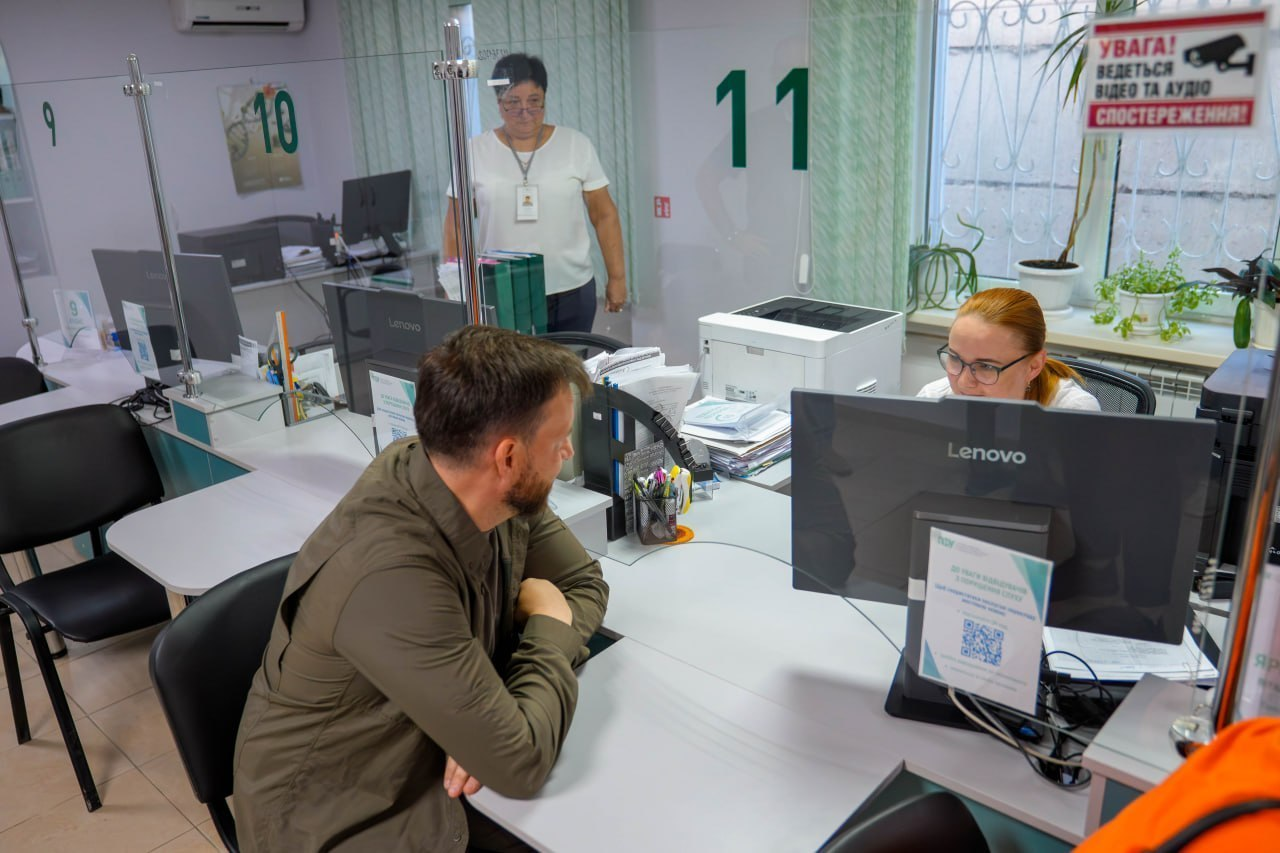 Робочий візит Міністра соціальної політики, сім’ї та єдності Дениса Улютіна до Головного управління Пенсійного фонду України в Херсонській області, де урядовець за робочим столом біля віконця обслуговування номер 11 спілкується з фахівчинею установи, яка адмініструє надання соціальних та пенсійних послуг громадянам в умовах воєнного стану.