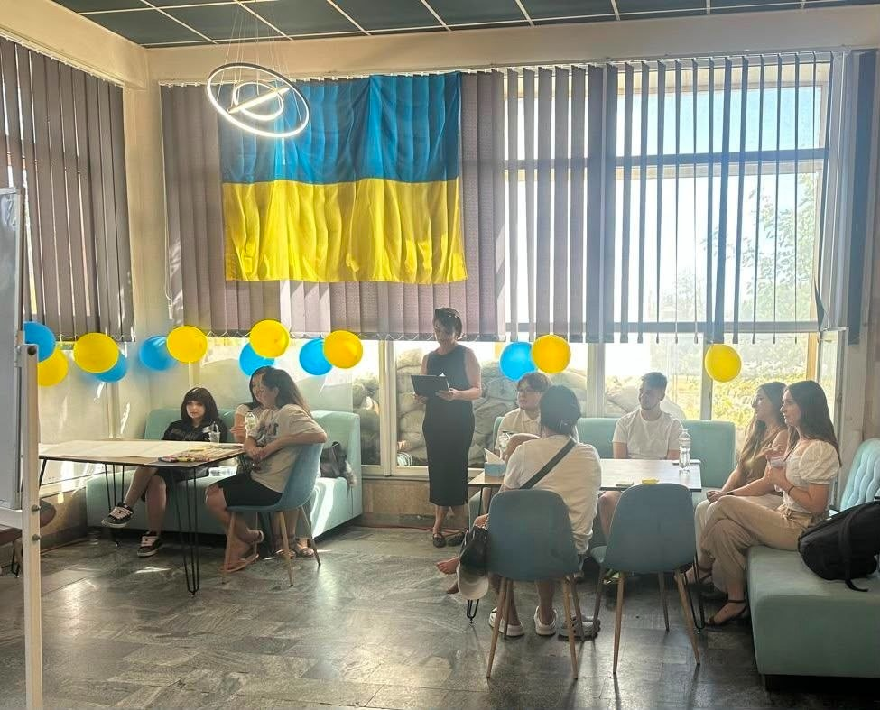 Молодь із числа ВПО з Херсонщини бере участь у командному флешмобі «Модель розвитку Херсонщини» в миколаївському координаційному центрі «Вільні разом». Учасники сидять на блакитних диванах і стільцях навколо столів, обговорюючи ідеї та проєкти відновлення рідного краю. Перед ними стоїть жінка-модератор у чорній сукні з текстом у руках. Приміщення прикрашене великим прапором України на вікні та ланцюжками з синіх і жовтих повітряних кульок.