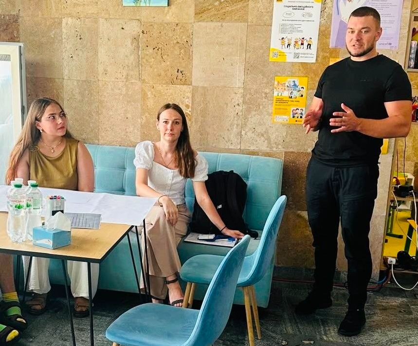 Учасники молодіжного заходу «Модель розвитку Херсонщини» в миколаївському координаційному центрі «Вільні разом» слухають виступ спікера. Чоловік у чорній футболці та спортивних штанях стоїть праворуч і активно жестикулює під час промови. Ліворуч на блакитному дивані за столом сидять дві дівчини, які уважно слухають доповідача та беруть участь у дискусії про майбутнє відновлення рідного краю. На столі перед ними лежать робочі матеріали, канцелярія та стоять пляшки з водою, а на стіні позаду висять інформаційні плакати.