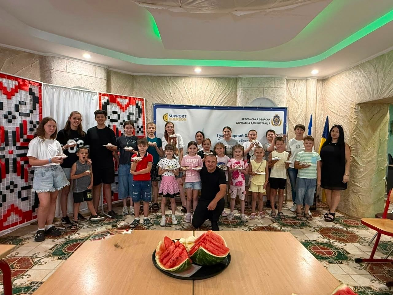 Спільне фото учасників дитячого майстер-класу з прикрашання кексів від координаційного центру «Вільні разом» та БФ «Сапорт Херсон» в Одесі. Велика група юних переселенців з Херсонщини та дорослих стоїть разом у залі на тлі офіційного банера «Гуманітарний штаб Херсонської обласної державної адміністрації / Support Kherson». Стіни приміщення прикрашені великими червоно-чорними орнаментами української вишиванки та державним прапором. Діти тримають у руках одноразові тарілки зі створеними власноруч кексами, а по центру перед ними присів один із організаторів. На передньому плані на дерев'яному столі стоїть велика тарілка зі стиглими скибками розрізаного херсонського кавуна, яким пригощали малечу.