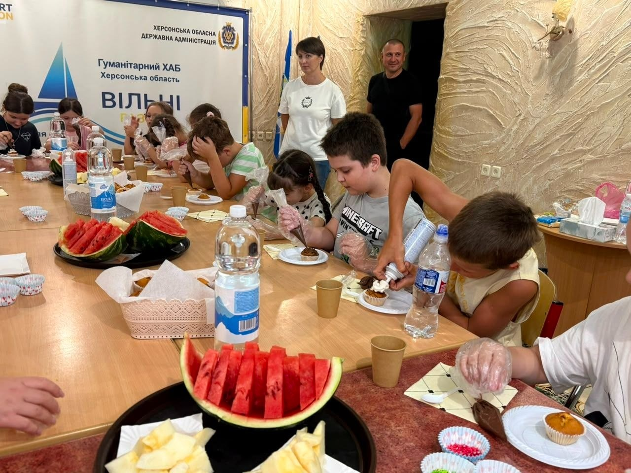 Процес прикрашання кексів на дитячому майстер-класі від координаційного центру «Вільні разом» та БФ «Сапорт Херсон» в Одесі. Юні переселенці з Херсонщини сидять за довгим дерев'яним столом у прозорих рукавичках, декоруючи власні смаколики за допомогою кондитерських мішків із кремом та кольорових посипок. На столі перед ними також стоять пляшки з водою, паперові стаканчики та великі тарілки зі стиглими шматочками херсонських кавунів і динь. На задньому плані за процесом спостерігають двоє дорослих організаторів, а ліворуч висить офіційний банер «Гуманітарний ХАБ Херсонська область Вільні разом».