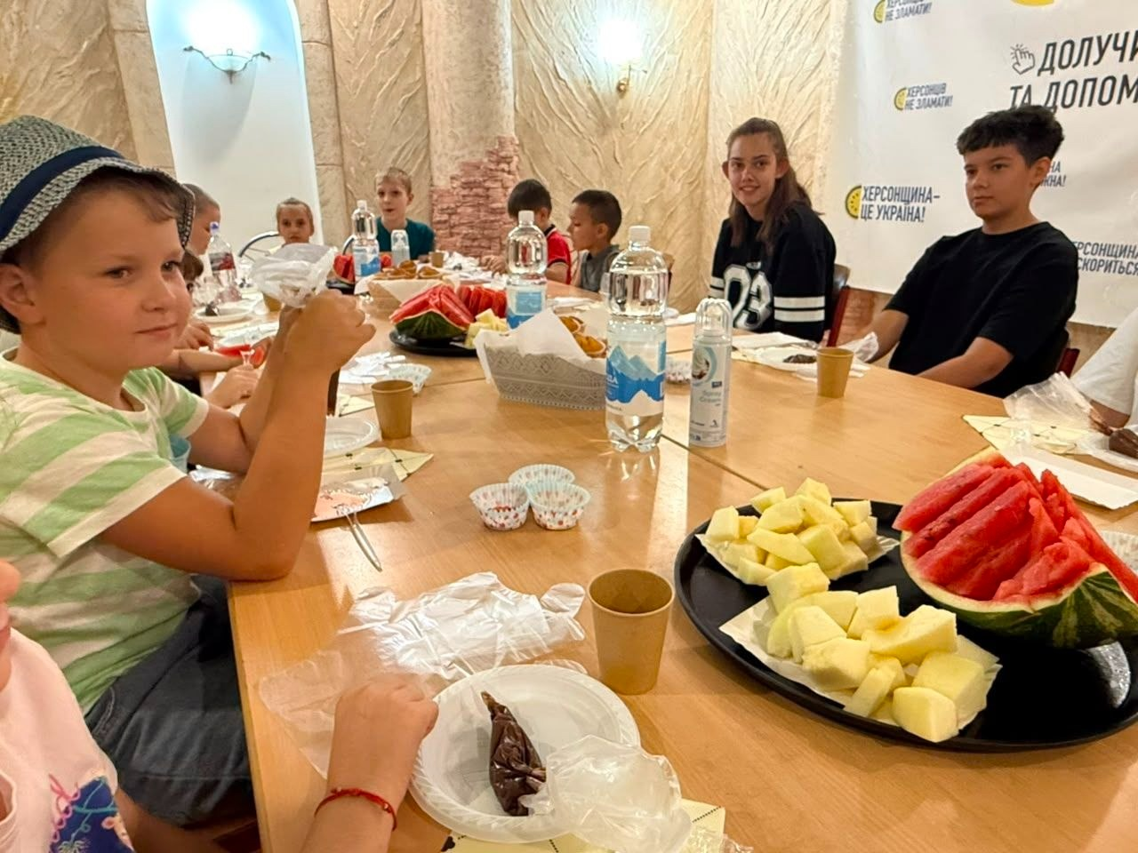 Юні учасники майстер-класу з прикрашання кексів від координаційного центру «Вільні разом» та БФ «Сапорт Херсон» в Одесі сидять за великим столом і готуються куштувати смаколики. Перед дітьми-переселенцями з Херсонщини лежать кондитерські мішки з кремом, паперові форми для випічки та серветки. На першому плані стоїть тарілка зі шматочками херсонського кавуна та дині, а також пляшки з водою та паперові стаканчики. На стіні праворуч розміщено банер із патріотичними написами: «Херсонщина — це Україна!» та «Долучайся та допоможи!».