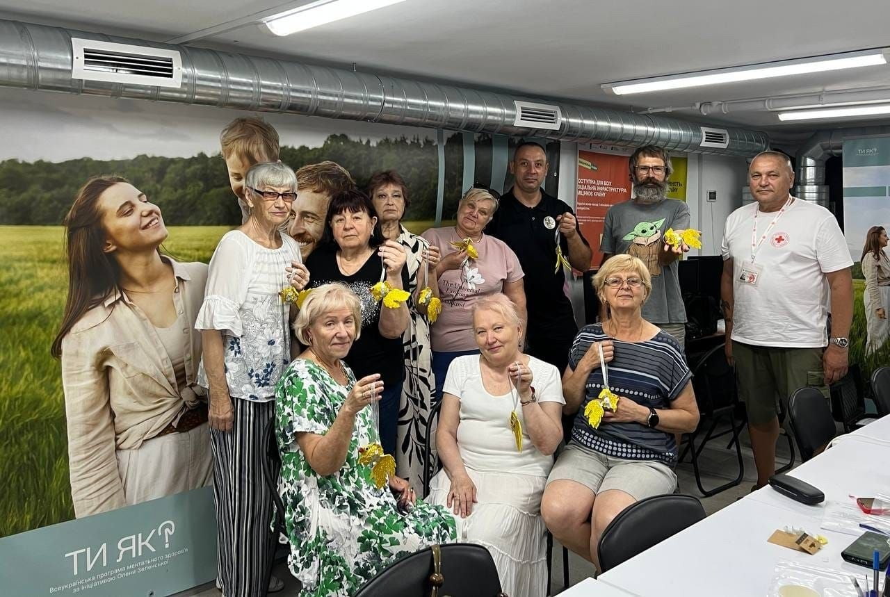 Спільне фото учасників та організаторів майстер-класу «Рибка бажання» в артмайстерні «Акваріум», що пройшов у Центрі життєстійкості. Група евакуйованих жінок старшого віку та волонтерів стоїть і сидить у приміщенні хабу, тримаючи в руках власноруч виготовлені блискучі жовті рибки-обереги на ниточках. Серед присутніх — представник Товариства Червоного Хреста України у білій фірмовій футболці та чоловік у сірій футболці з малюнком малюка Йоди. Ліворуч на стіні розміщено великий зелений банер Всеукраїнської програми ментального здоров'я «Ти як?» за ініціативою Олени Зеленської, на тлі якого фотографуються учасники події.