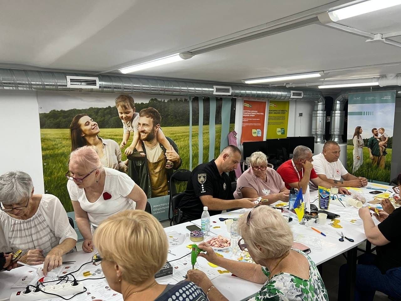 Процес виготовлення поробок на майстер-класі «Рибка бажання» в артмайстерні «Акваріум», що пройшов у межах тижня психосоціальної підтримки в Центрі життєстійкості. Учасники переважно старшого віку сидять за довгим білим столом, зосереджено займаючись рукоділлям. По центру разом із ними працює чоловік у чорній формі Поліції охорони, а праворуч за столом сидить волонтер Товариства Червоного Хреста України у білій фірмовій футболці. На столі розкладено клейові пістолети, кольоровий папір, ножиці та заготовки для оберегів. На стінах кімнати розміщено великі інформаційні банери Міністерства соціальної політики України та Всеукраїнської програми ментального здоров'я «Ти як?», які підкреслюють мету заходу — стабілізацію стану та зняття напруги через творчість.