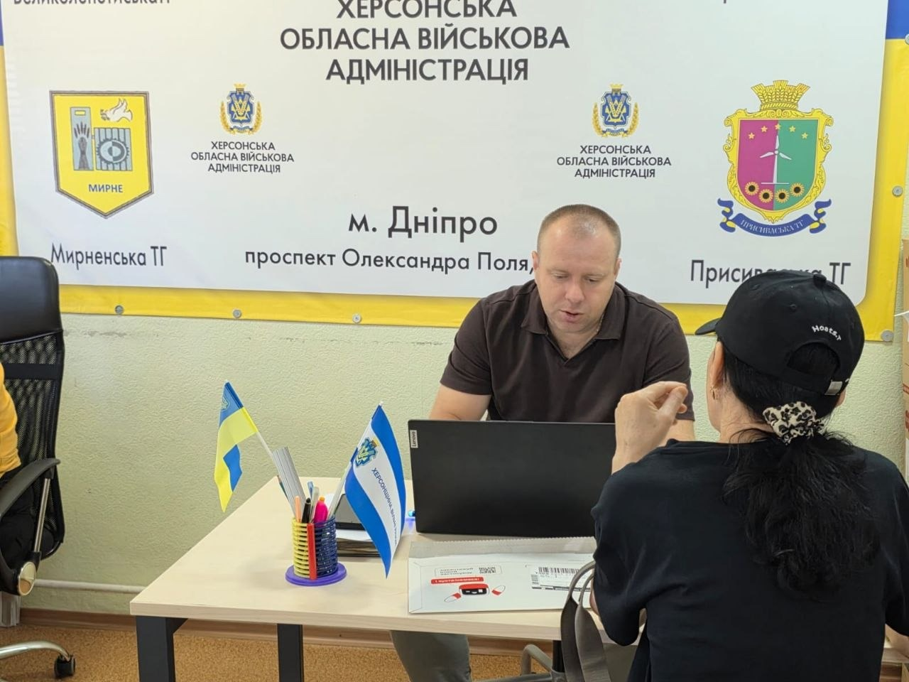 Індивідуальна консультація переселенки заступником начальника Управління Пенсійного фонду в Херсонській області Олегом Чабановим у дніпровському центрі «Вільні разом». Посадовець у коричневій футболці поло сидить за столом із ноутбуком, роз'яснюючи жінці в чорній кепці питання пенсій та стажу в межах платформи «Діалог з владою». На столі стоять настільні прапорці України та Херсонщини, канцелярія та документи. Позаду на стіні розміщено офіційний банер Херсонської ОВА з гербами Мирненської і Присиваської громад та адресою на проспекті Олександра Поля у Дніпрі.