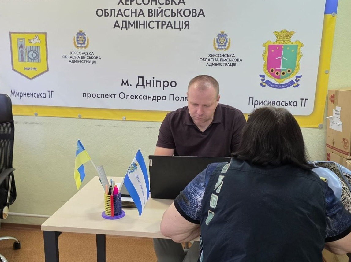 Індивідуальна консультація переселенки заступником начальника Управління Пенсійного фонду в Херсонській області Олегом Чабановим у дніпровському центрі «Вільні разом». Посадовець у коричневій футболці поло сидить за столом із ноутбуком, консультуючи жінку в синій футболці з візерунком. На столі стоять настільні прапорці України та Херсонщини й канцелярія. Позаду на стіні розміщено офіційний банер Херсонської ОВА з гербами Мирненської і Присиваської громад та адресою у Дніпрі.