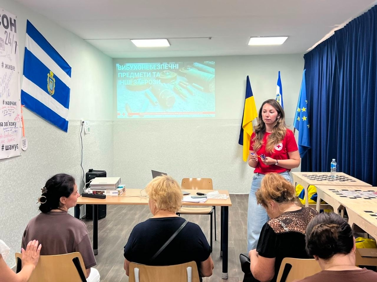 нформаційний захід з техніки мінної безпеки для ВПО з Херсонщини в хмельницькому координаційному центрі «Вільні разом». Представниця Хмельницької обласної організації Товариства Червоного Хреста України у червоній фірмовій футболці проводить сесію «Інформування про ризики вибухонебезпечних предметів та правила безпечнішої поведінки». Позаду лекторки увімкнено проєктор із презентацією «Вибухонебезпечні предмети та інші загрози», поруч стоять прапори України та ЄС. У залі на стільцях сидять слухачі, які вивчають алгоритм дій у разі виявлення підозрілих знахідок. Ліворуч на стіні висить прапор із гербом міста Херсон.