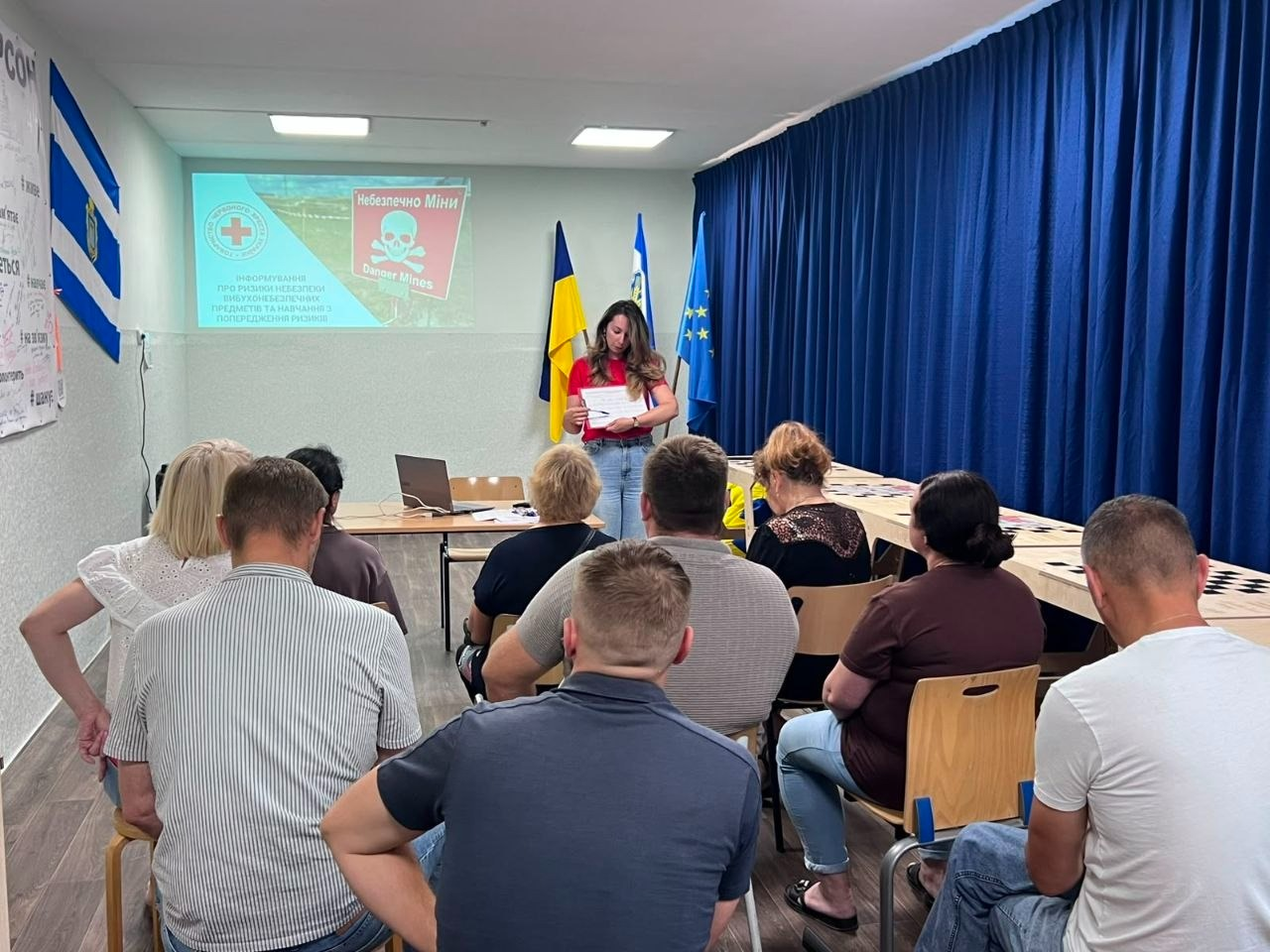 Інформаційний захід з техніки мінної безпеки для ВПО з Херсонщини в хмельницькому координаційному центрі «Вільні разом». Представниця Хмельницької обласної організації Товариства Червоного Хреста України у червоній футболці тримає інформаційні матеріали, виступаючи перед слухачами. Позаду неї увімкнено проєктор зі слайдом «Небезпечно Міни / Danger Mines» та логотипом організації. У залі на стільцях спиною до камери сидять учасники тренінгу, які вивчають правила безпечної поведінки. Локація прикрашена прапорами України, ЄС та Херсонщини, а ліворуч на стіні висить прапор із гербом міста Херсон.