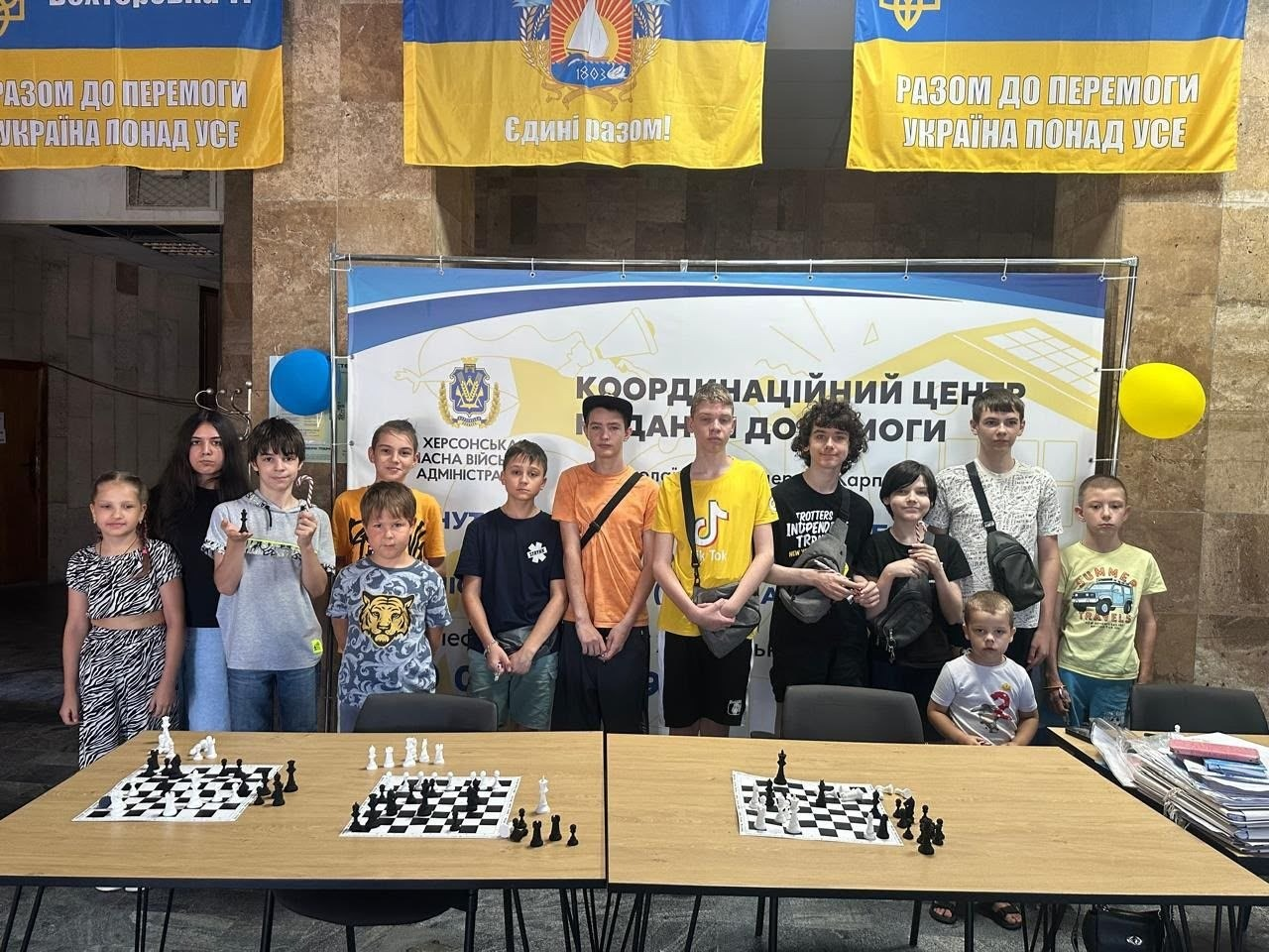 Спільне фото учасників дитячого шахового турніру для юних переселенців з Херсонщини в миколаївському центрі «Вільні разом». Діти та підлітки стоять за столами з розставленими шаховими дошками на тлі банера Херсонської ОВА «Координаційний центр». Зал прикрашений синьо-жовтими прапорами з гаслами «Разом до перемоги» та повітряними кульками.