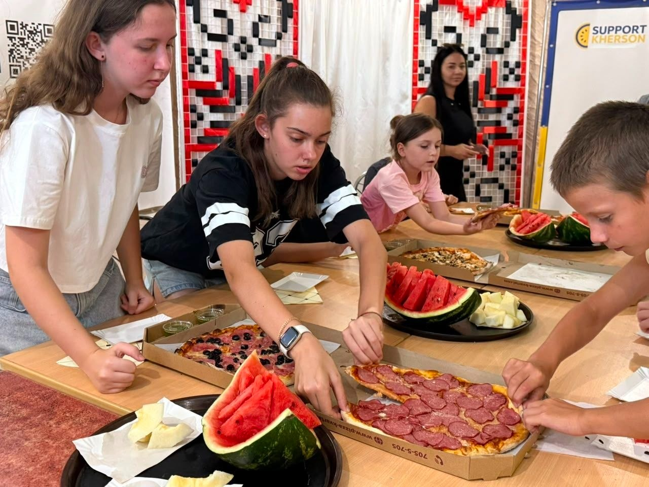 Юні переселенці з Херсонщини пригощаються піцою від ресторану «Італійський квартал» під час майстер-класу з 3D-друку від майстерні «Мудрі» та БФ «Сапорт Херсон» у координаційному центрі. Діти та підлітки зібралися навколо великого дерев'яного столу, беручи шматочки піци з відкритих картонних коробок. На столі також стоять тарілки зі стиглими скибками херсонського кавуна та дині. На задньому плані видно настінні панно з червоно-чорним орнаментом української вишиванки, QR-код та край офіційного банера «Support Kherson».