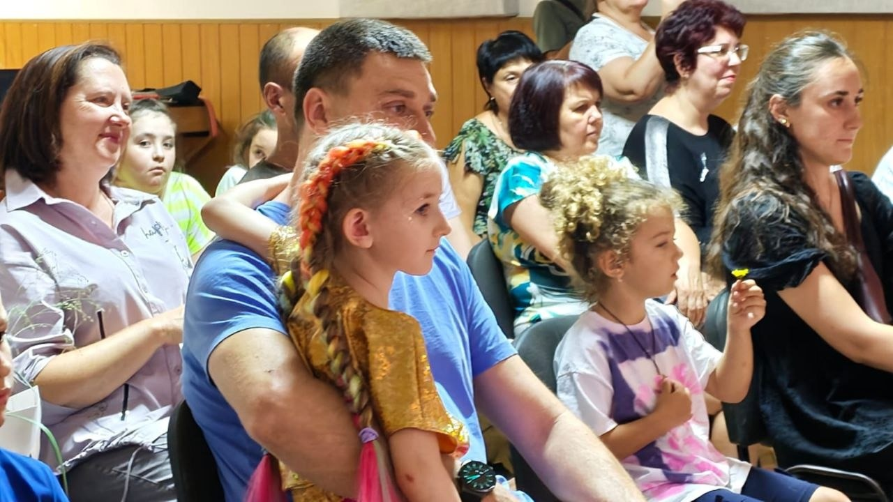 Глядачі в залі під час звітного концерту вокальної студії Херсонського обласного палацу культури. На передньому плані чоловік у синій футболці тримає на колінах дівчинку в блискучій золотистій сукні з яскравими кольоровими канекалонами у косах. Поруч сидить маленька кучерява дівчинка у різнокольоровій футболці тай-дай, яка тримає маленьку жовту квітку, а праворуч від неї — молода жінка з довгим темним волоссям. На задньому плані на стільцях сидять інші глядачі, переважно жінки, які уважно й емоційно спостерігають за виступами юних вокалістів студії Еріки Ситник.