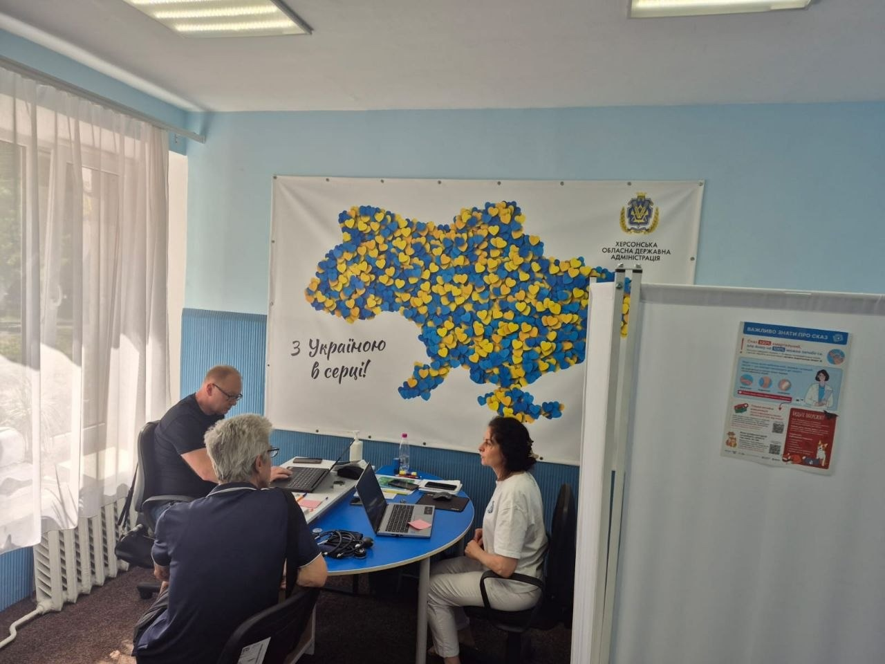 День медичних консультацій для переселенців з Херсонщини від лікарки-терапевтки з Маріуполя за підтримки Червоного Хреста у хмельницькому центрі «Вільні разом». За круглим блакитним столом пацієнтка отримує індивідуальні рекомендації від двох фахівців, які працюють за ноутбуками. Прийом проходить на тлі банера Херсонської ОВА з картою України із синьо-жовтих сердечок та написом «З Україною в серці!».