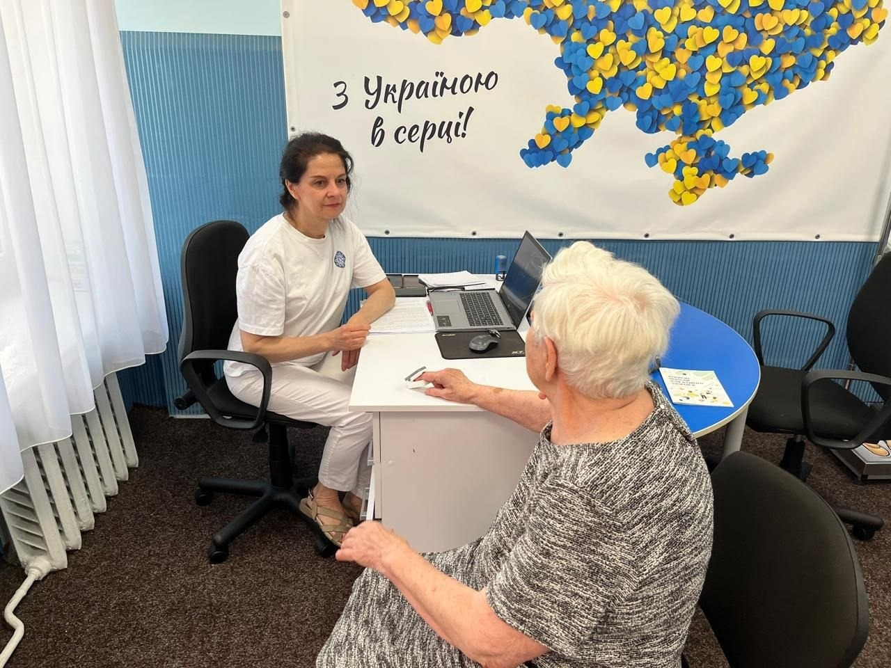 Медична консультація для ВПО з Херсонщини від лікарки-терапевтки ЦПМСД № 3 Маріуполя за підтримки Червоного Хреста у хмельницькому центрі «Вільні разом». Лікарка у світлій медичній формі сидить за білим столом із ноутбуком, консультуючи літню пацієнтку щодо результатів скринінгу та лікування. Прийом проходить на тлі офіційного банера з картою України із синьо-жовтих сердечок та написом «З Україною в серці!».