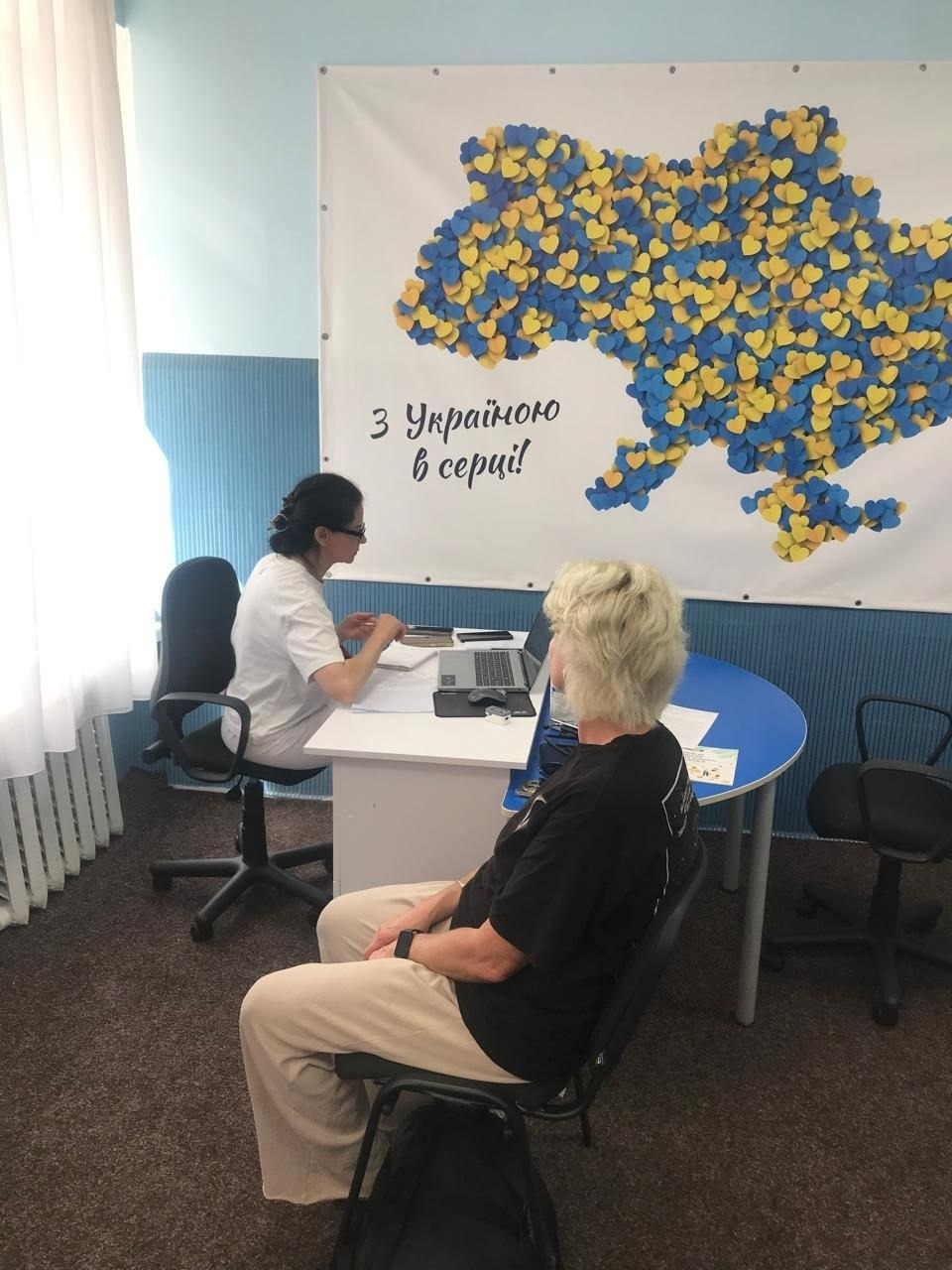Медична консультація для ВПО з Херсонщини від лікарки-терапевтки ЦПМСД № 3 Маріуполя за підтримки Червоного Хреста у хмельницькому центрі «Вільні разом». Лікарка у світлій медичній формі сидить за столом із ноутбуком, консультуючи жінку в чорній футболці та світлих штанах у межах програми «40+». Прийом проходить на тлі банера з картою України із синьо-жовтих сердечок та написом «З Україною в серці!».