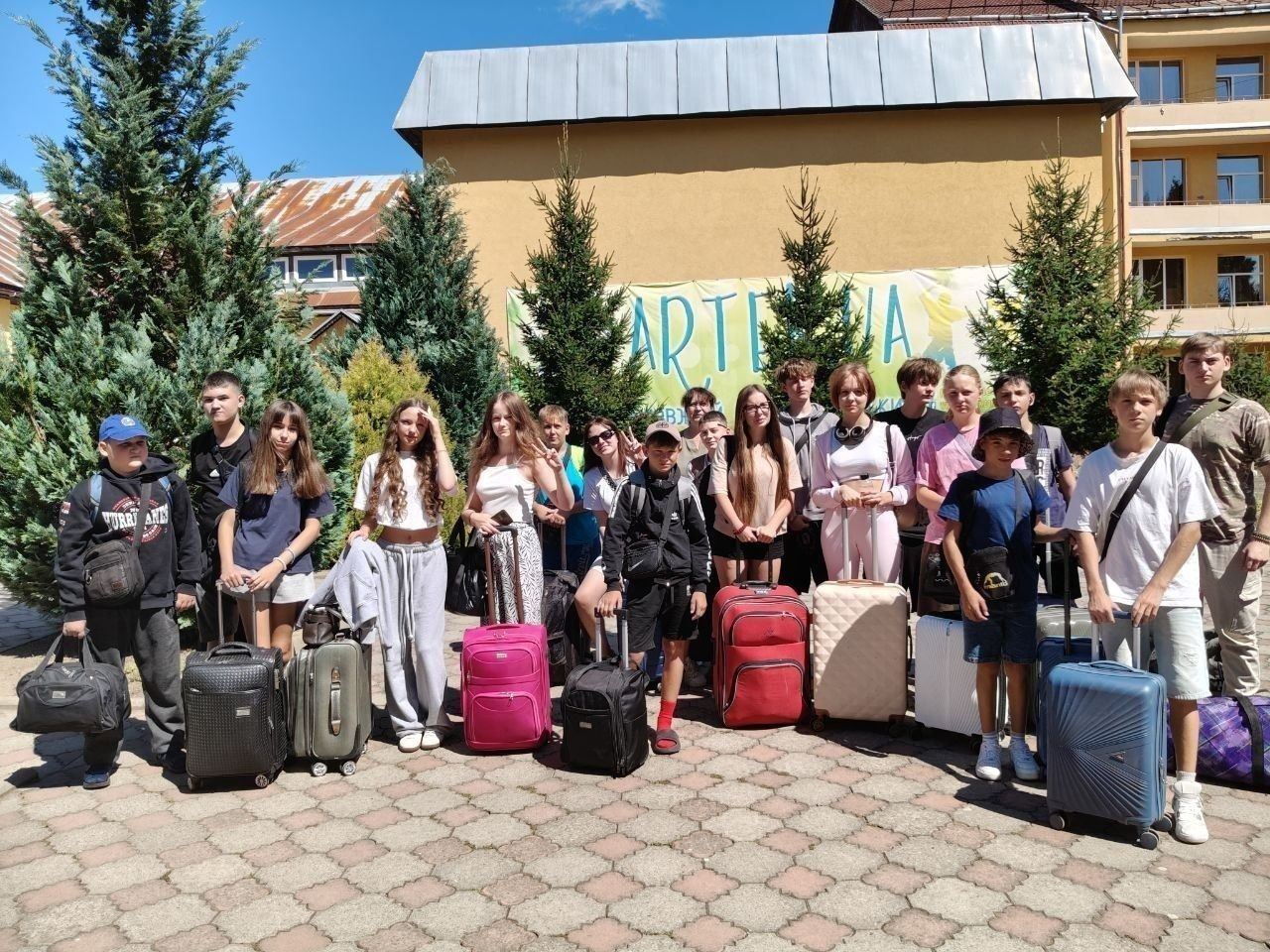 Група дітей та підлітків з-поміж ВПО Херсонщини позує для спільного фото на брущатому подвір’ї Міжнародного дитячого центру «Артек» перед початком оздоровчої зміни. Хлопці та дівчата різного віку тримають у руках або тримають перед собою великі дорожні валізи (рожеві, червоні, чорні, сині) та наплічники. Вони вдягнені у літній повсякденний одяг: футболки, кепки, шорти та спортивні штани. Локація розташована під яскравим сонцем на тлі високих вічнозелених ялин та двоповерхового жовтого корпусу з коричневим дахом. Позаду учасників на стіні будівлі розтягнуто банер із великим розмитим написом «ARTEK.UA», а сама поїздка організована за сприяння Департаменту соціального розвитку Херсонської ОДА.