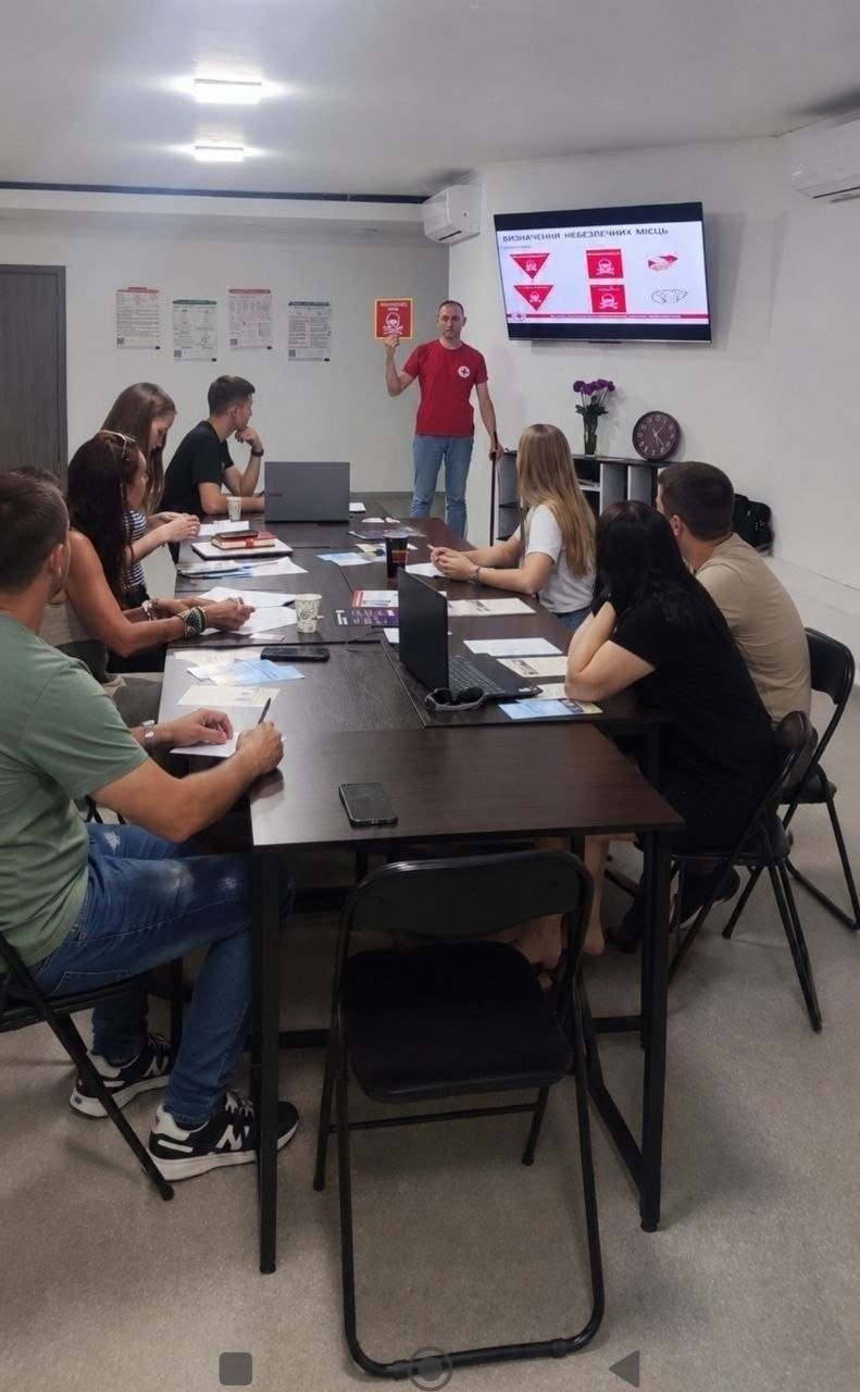 Лектор у червоній футболці з логотипом Червоного Хреста проводить для переселенців навчання з мінної безпеки, організоване Координаційним центром «Вільні разом» та ГО «Сталеві духом» у Черкасах. Учасники сидять за довгим столом у приміщенні центру в Черкасах, слухають спікера, який тримає попереджувальну картку, та дивляться на екран із презентацією «Визначення небезпечних місць».