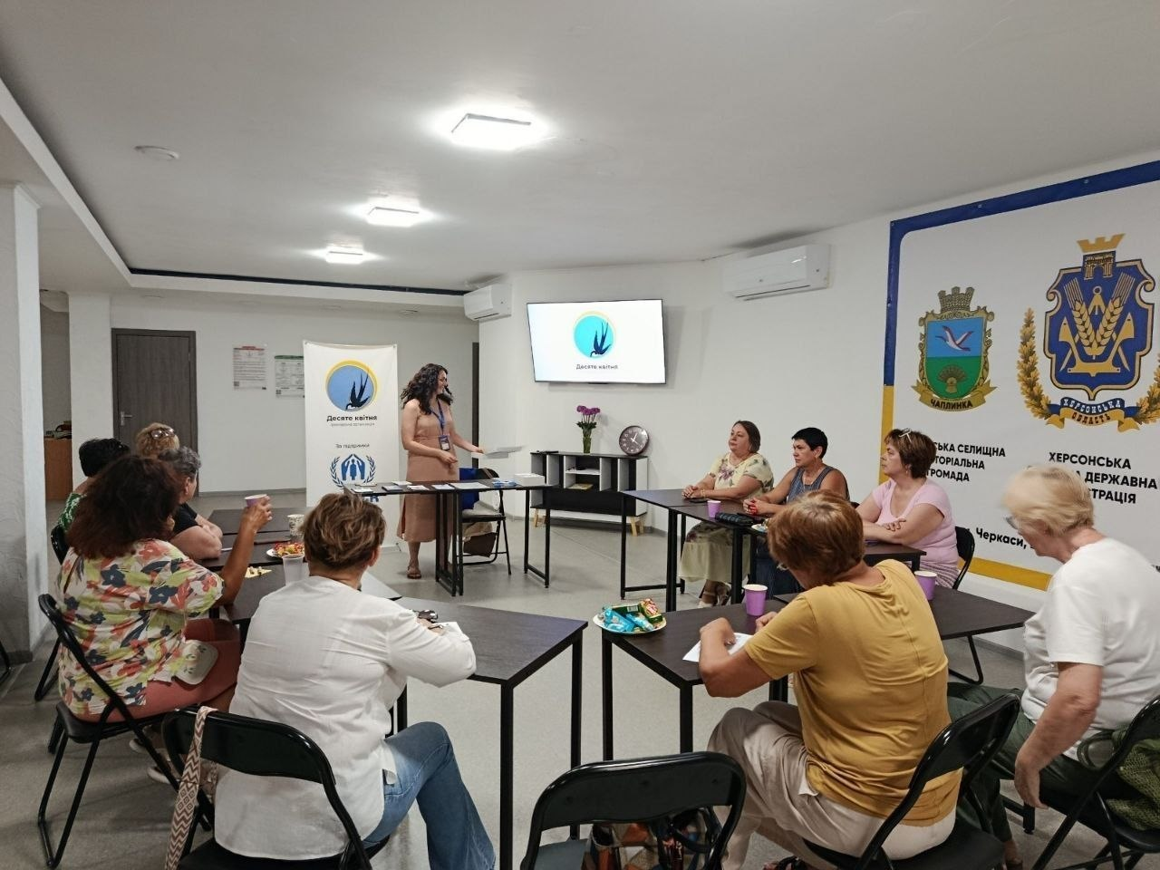 Спікерка проводить лекцію-обговорення «Червоні прапорці в стосунках» для групи жінок-переселенок із Херсонщини в Координаційному центрі «Вільні разом» у Черкасах. На задньому плані видно екран та ролап із логотипом ГО «Десяте квітня», а на стіні праворуч — великий банер із символікою Чаплинської громади та Херсонської ОДА.