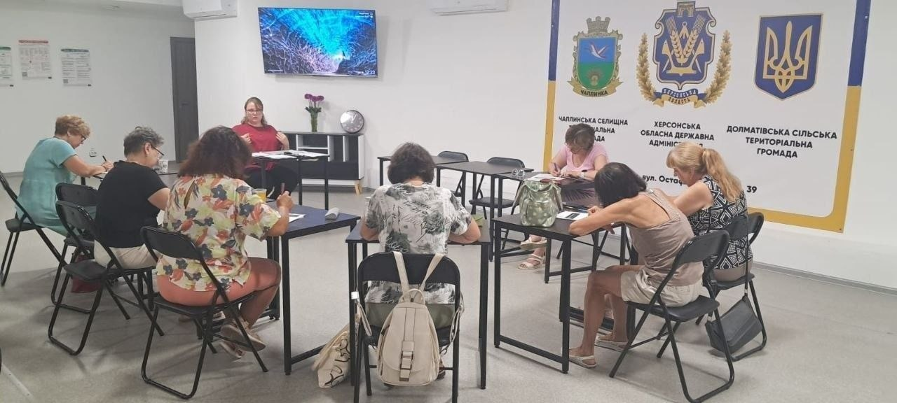 Група жінок-переселенок із Херсонщини сидить за столами у світлій залі під час заняття з англійської мови, організованого Координаційним центром «Вільні разом» у Черкасах. Учасниці роблять записи у зошитах, а попереду перед ними виступає викладачка. Праворуч на стіні розміщено великий інформаційний банер із гербами Чаплинської селищної, Долматівської сільської громад та Херсонської ОДА.