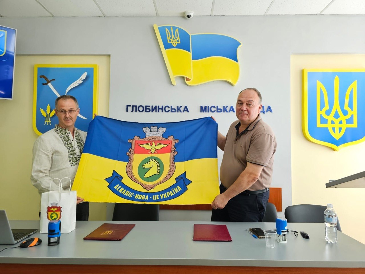 Два представники Глобинської та Асканіє-Нова громад стоять у залі засідань Глобинської міської ради й тримають синьо-жовтий прапор. У центрі полотнища зображено герб Асканії-Нови із золотим орлом та головою коня тарпана, а знизу розміщено стрічку з написом «Асканія-Нова — це Україна». На стіні позаду них міститься напис «Глобинська міська рада», державна символіка України та офіційний герб Глобинської громади.