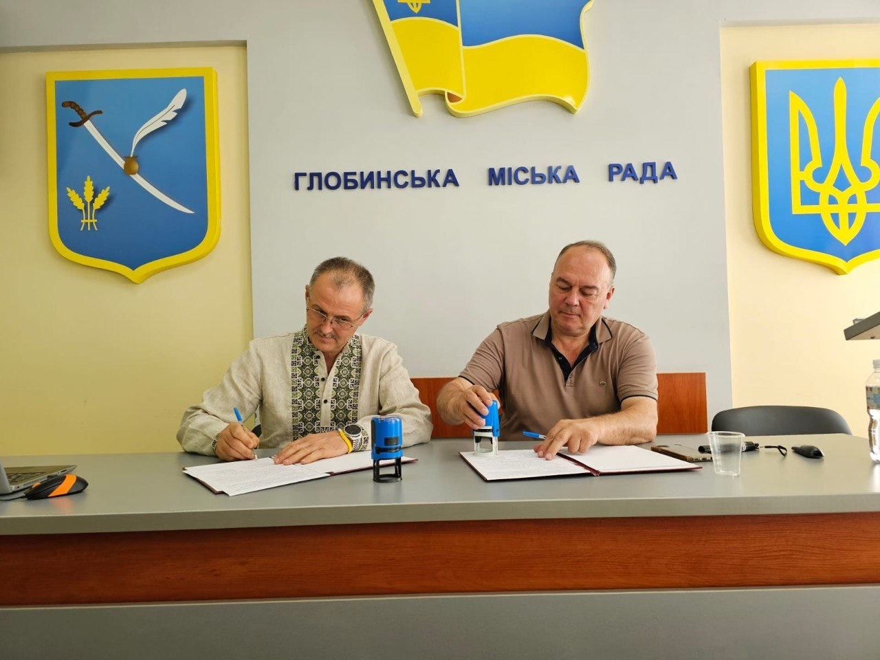 Представники Глобинської міської ради (Полтавщина) та Асканіє-Нова селищної громади (Херсонщина) сидять за столом у залі засідань і підписують Меморандум про співпрацю в межах проєкту «Пліч-о-пліч». На столі лежать відкриті теки з документами та канцелярські печатки, а на білій стіні позаду них розміщено напис «Глобинська міська рада», малий герб і прапор України та офіційний герб Глобинської громади.