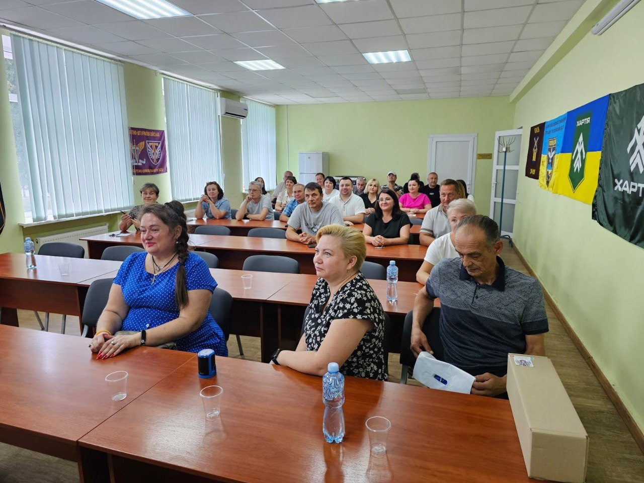 Представники та мешканці Асканіє-Нова селищної громади (Херсонщина) та Глобинської міської громади (Полтавщина) сидять в аудиторії під час зустрічі щодо підписання Меморандуму про співпрацю в межах проєкту «Пліч-о-пліч». На передньому плані за дерев'яним столом сидить керівництво громад, а на стіні праворуч розміщені прапори військових підрозділів, зокрема 13-ї бригади НГУ «Хартія» та 3-ї ОШБр.