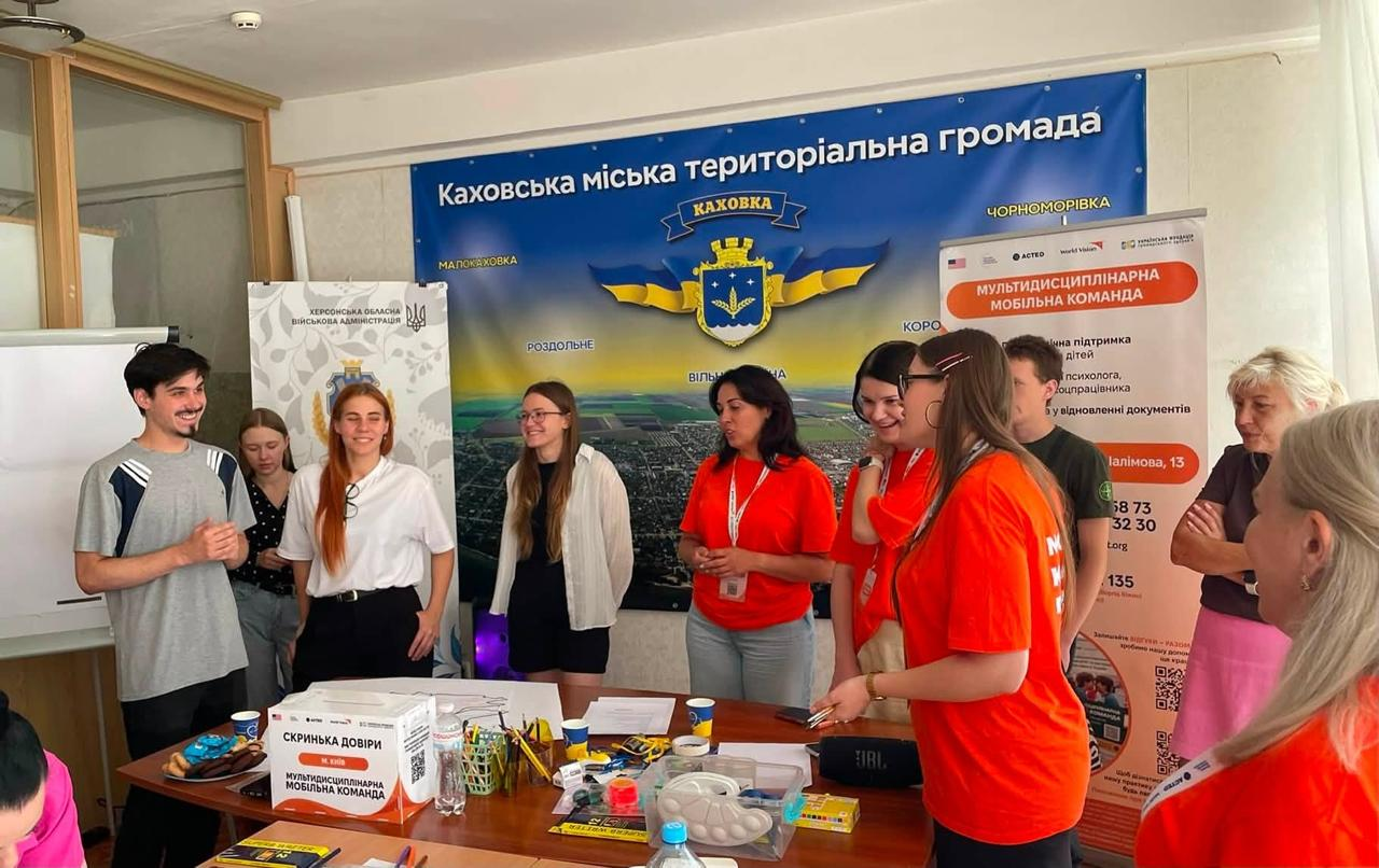Учасники молодіжного флешмобу «Модель розвитку Херсонщини» — представники ГО «Молодіжний конгрес Херсонщини» та Каховської міської громади — зібралися навколо робочого столу в київському центрі «Вільні разом». На тлі офіційного банера громади та стенда Мультидисциплінарної мобільної команди молодь в яскравих помаранчевих футболках організації, повсякденних сорочках, футболках і блузах активно обговорює плани відновлення регіону, використовуючи розкладену на столі канцелярію та «Скриньку довіри».