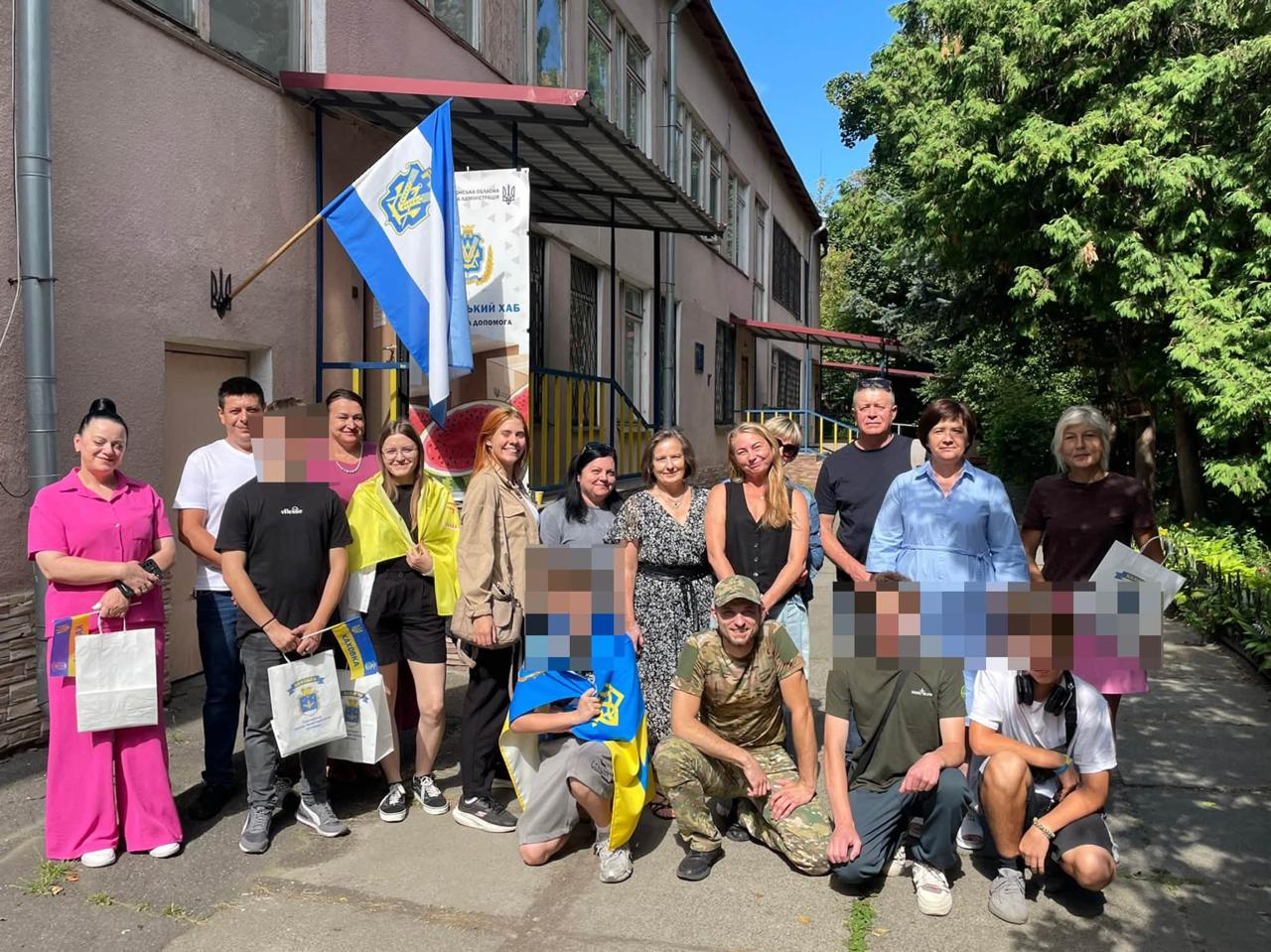 Група учасників та організаторів молодіжного флешмобу «Модель розвитку Херсонщини» позує для спільного фото на вулиці біля входу до центру допомоги «Вільні разом» у Києві. На тлі будівлі хабу майорить синьо-білий прапор Каховки та встановлено офіційний банер ХОДА, а присутні цивільні особи, підлітки та військовослужбовець у камуфляжі тримають святкові паперові пакети, прапорці й символіку громади.