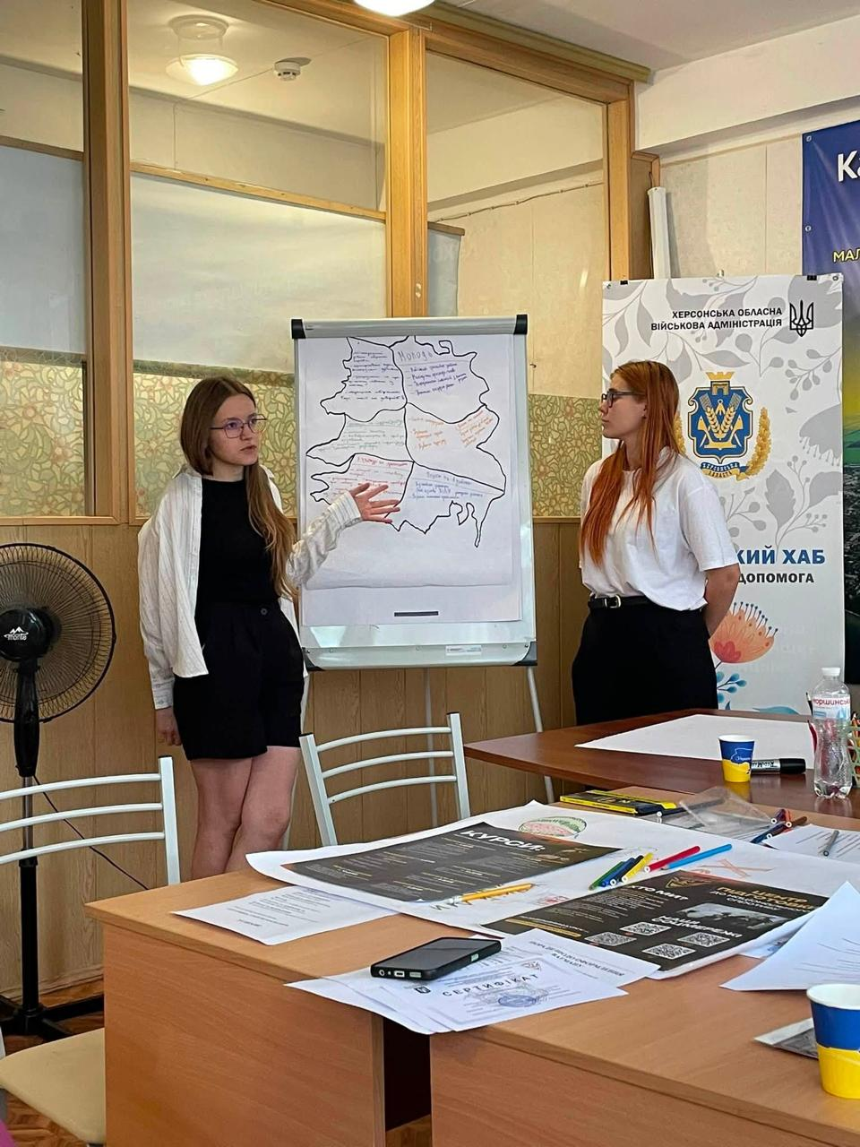 Дві учасниці молодіжного флешмобу «Модель розвитку Херсонщини» презентують напрацьовану мапу регіону під час командної роботи в київському центрі «Вільні разом». Русява дівчина в окулярах, білій сорочці поверх чорного топа та чорних шортах жестом показує на закріплений на фліпчарті великий аркуш із намальованими контурами карти та текстовими нотатками, тоді як поруч стоїть рудоволоса учасниця в білій сорочці та чорних штанах на тлі офіційного банера Херсонського хабу.