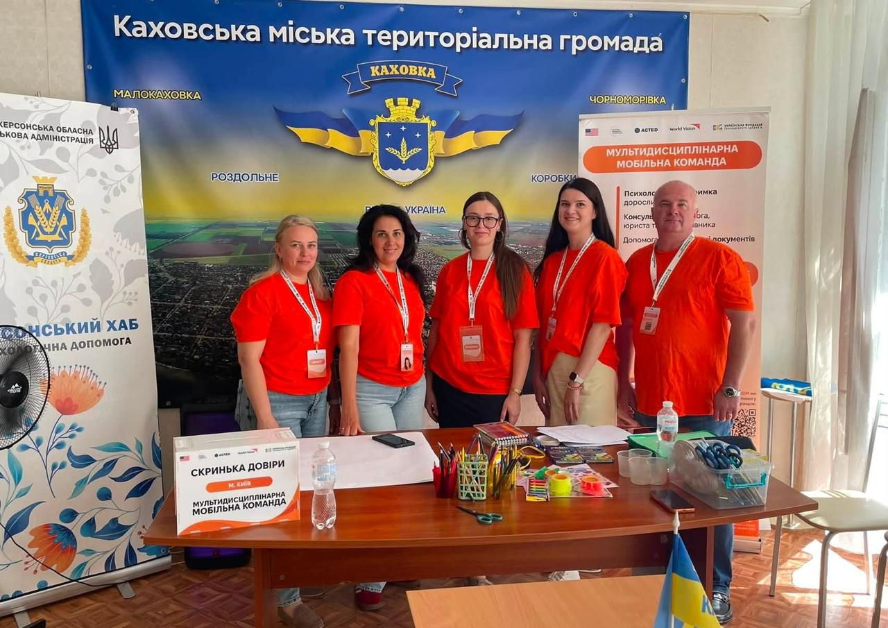 Представники Мультидисциплінарної мобільної команди позують для спільного фото в київському центрі «Вільні разом» під час молодіжного флешмобу «Модель розвитку Херсонщини». Чотири жінки з русявим, темним та довгим каштановим волоссям, а також один чоловік одягнені в однакові брендовані яскраві помаранчеві футболки з білими бейджами на шиї. Вони стоять біля робочого столу із канцелярією та білою «Скринькою довіри» на тлі великого офіційного банера «Каховська міська територіальна громада» та інформаційного стенда організації.