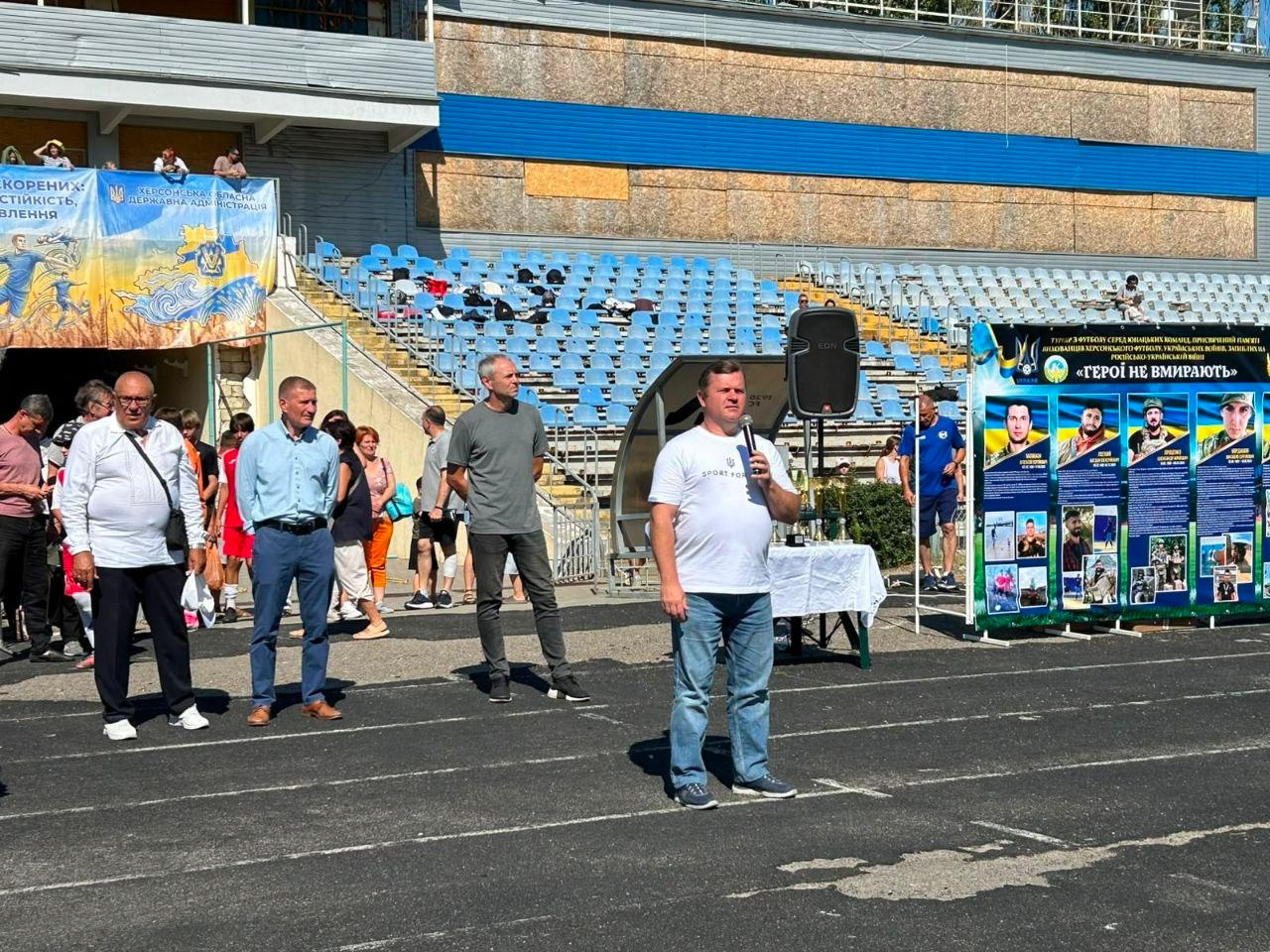 Заступник голови Херсонської ОДА Володимир Клюцевський у білій футболці та джинсах говорить у мікрофон на відкритті футбольного турніру «ГЕРОЇ НЕ ВМИРАЮТЬ» на стадіоні в Миколаєві. Позаду нього лівіше, серед двох інших чоловіків, стоїть директор Херсонського фахового спортивного коледжу Валентин Сух, одягнений у світло-блакитну сорочку та сині штани. На передньому плані видно бігову доріжку, а на тлі розташовані трибуни з синіми сидіннями, банер Херсонської ОДА та великий інформаційний стенд із портретами загиблих військовослужбовців-футболістів.