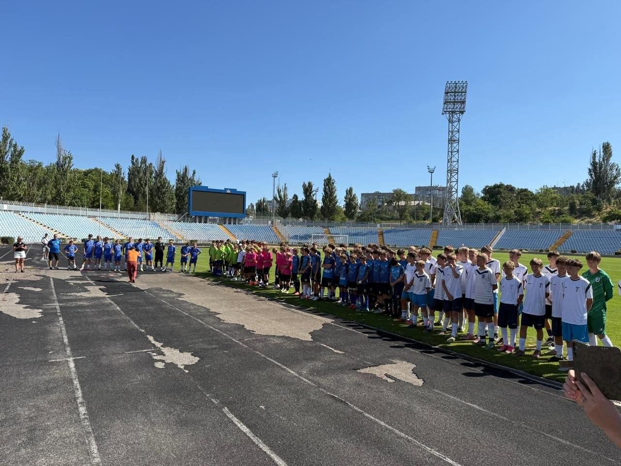Юнацькі футбольні команди, які взяли участь у відкритому Херсонському обласному турнірі «ГЕРОЇ НЕ ВМИРАЮТЬ» 2026, вишикувалися в шеренгу на полі стадіону в Миколаєві. Спортсмени у різноколірній формі (білій, синій, рожевій, зеленій) стоять уздовж асфальтованої бігової доріжки, де ліворуч також видно організаторів та гостей заходу. На задньому плані, під ясним синім небом, розташовані великі трибуни з блакитними сидіннями, велике електронне табло стадіону та металева щогла нічного освітлення.