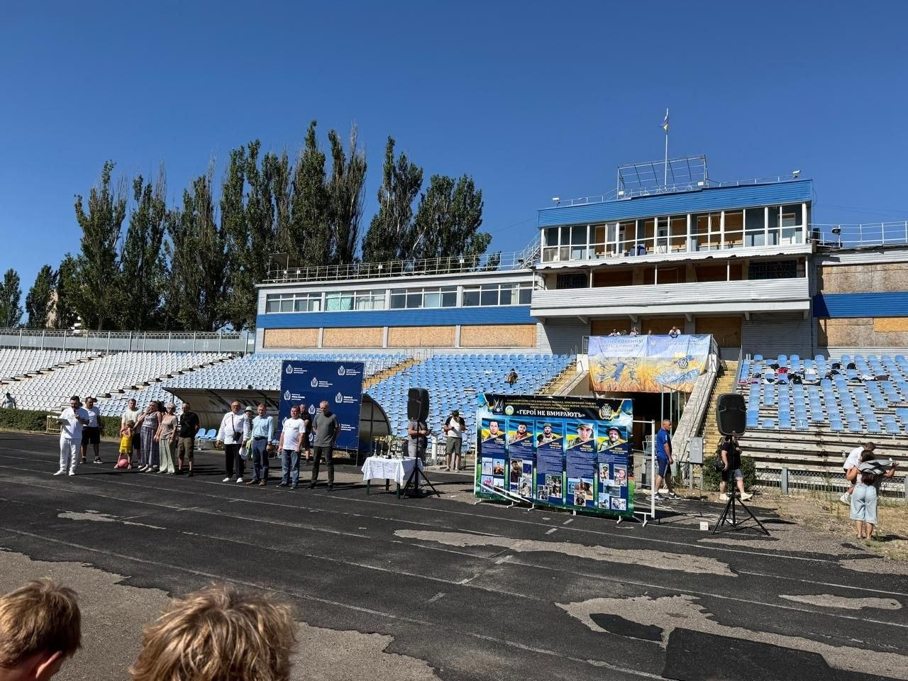 На біговій доріжці стадіону в Миколаєві під час футбольного турніру «ГЕРОЇ НЕ ВМИРАЮТЬ» вишикувалися в шеренгу офіційні гості та організатори заходу. У лівій частині кадру заступник директора Департаменту реалізації гуманітарної політики Херсонської ОДА Андрій Єрьоменко у білій футболці та білих штанах говорить у мікрофон, тримаючи в руці аркуш із промовою. Поруч із ним у шерензі стоїть директор Херсонського фахового спортивного коледжу Валентин Сух у світло-блакитній сорочці та синіх штанах, а праворуч від нього — заступник голови Херсонської ОДА Володимир Клюцевський у білій футболці та синіх джинсах. У правій частині розташований великий інформаційний банер із портретами п'яти загиблих військовослужбовців-вихованців херсонського футболу, а поруч стоїть стіл із кубками. На задньому плані, під ясним небом, підіймаються трибуни стадіону з блакитними сидіннями та височіє двоповерхова будівля коментаторської вишки з флагштоком.