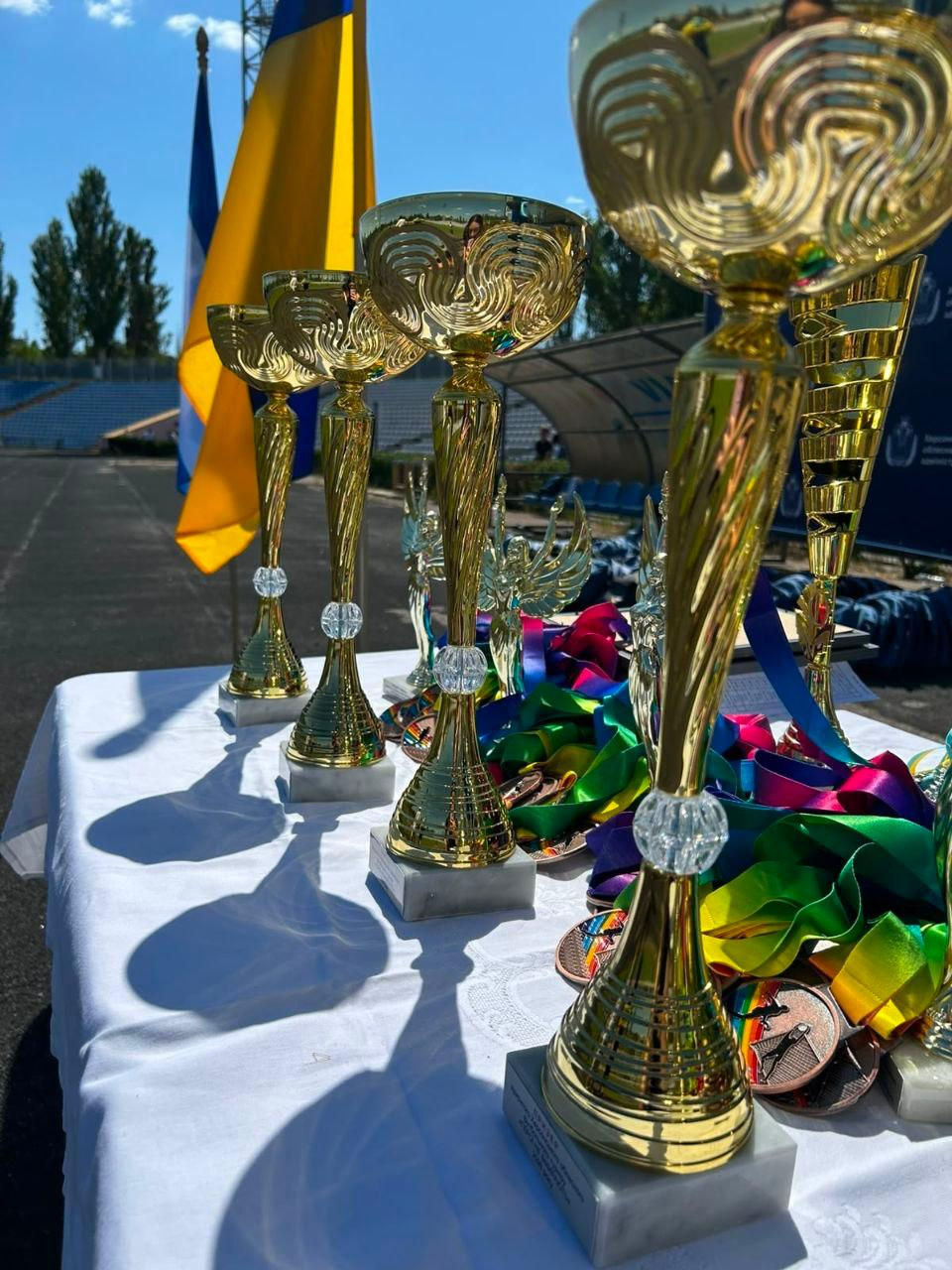 На передньому плані білого столу крупним планом вишикувані в ряд великі золоті кубки на мармурових підставках для призерів футбольного турніру «ГЕРОЇ НЕ ВМИРАЮТЬ». Поруч із нагородами розкладені медалі з яскравими різноколірними та синьо-жовтими стрічками. На розмитому задньому плані під синім небом видно державний прапор України, що майорить на вітрі біля бігових доріжок стадіону в Миколаєві.