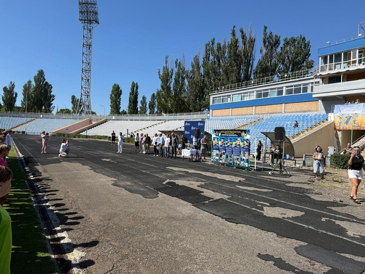 На біговій доріжці стадіону в Миколаєві під час футбольного турніру «ГЕРОЇ НЕ ВМИРАЮТЬ» вишикувалися в шеренгу офіційні гості та організатори заходу. У лівій частині кадру заступник директора Департаменту реалізації гуманітарної політики Херсонської ОДА Андрій Єрьоменко у білій футболці та білих штанах говорить у мікрофон, тримаючи в руці аркуш із промовою. Поруч із ним у шерензі стоїть директор Херсонського фахового спортивного коледжу Валентин Сух у світло-блакитній сорочці та синіх штанах, а праворуч від нього — заступник голови Херсонської ОДА Володимир Клюцевський у білій футболці та синіх джинсах. У правій частині розташований великий інформаційний банер із портретами п'яти загиблих військовослужбовців-вихованців херсонського футболу, а поруч стоїть стіл із кубками. На передньому плані зліва дівчина-фотограф присіла, щоб зробити знімок. На задньому плані, під ясним небом, підіймаються трибуни стадіону з блакитними сидіннями, височіє металева щогла нічного освітлення та двоповерхова будівля коментаторської вишки з флагштоком.