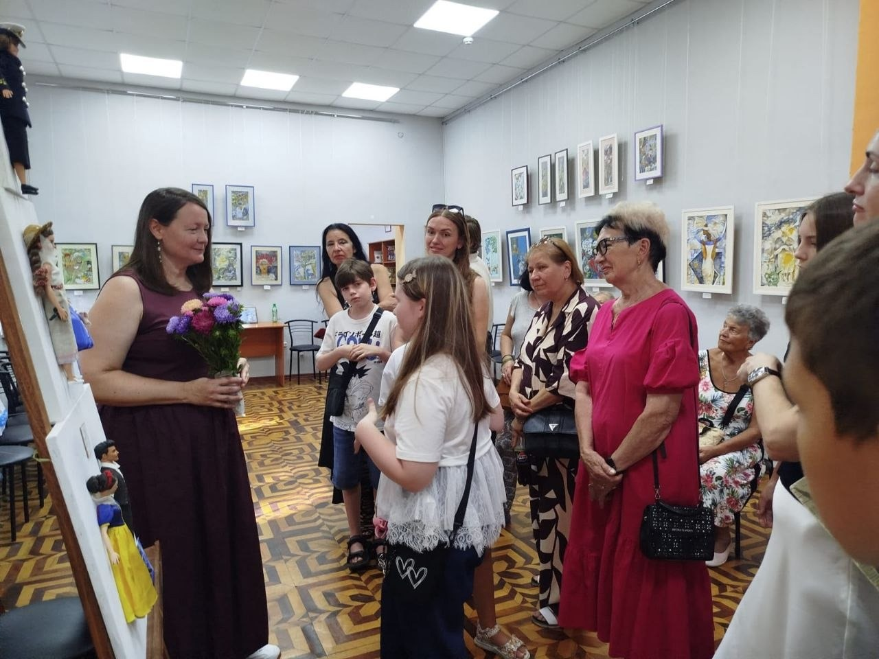 Група переселенців з Херсонщини та представники хабу «Вільні разом» на виставці авторських ляльок проєкту «КуклЬтура» в Одеській бібліотеці уважно слухають колекціонерку Олену Філіппову, яка тримає букет квітів на тлі виставкових стендів та картин.