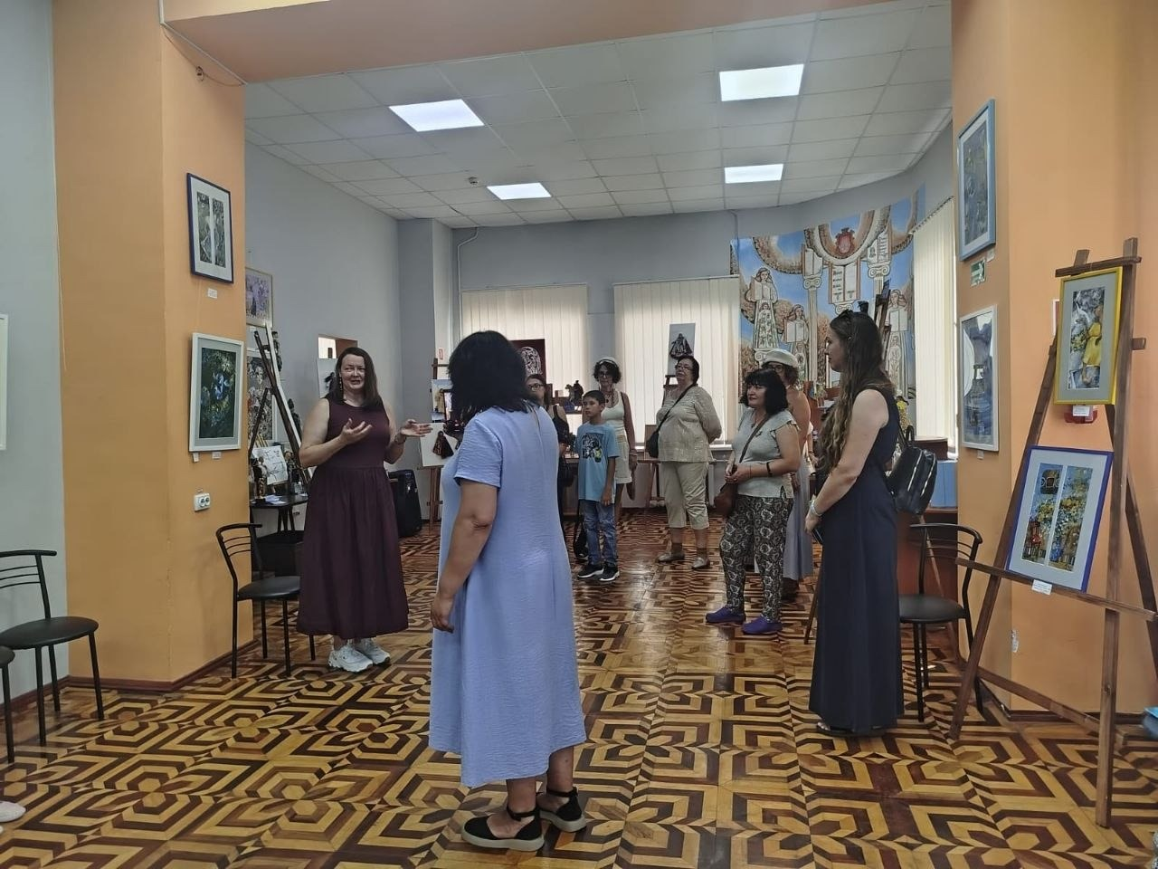 Колекціонерка Олена Філіппова у бордовій сукні проводить екскурсію виставкою «КуклЬтура» для групи переселенців з Херсонщини у великій залі Одеської бібліотеки. Відвідувачі у літньому одязі стоять півколом та слухають розповідь на тлі розвішаних на стінах картин і виставкових стендів.